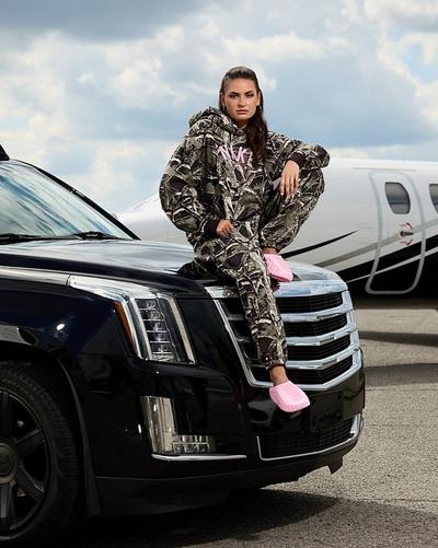 Person wearing a camouflage Nike hoodie and matching pants with bright pink slides sits on the hood of a black SUV beside a private plane.