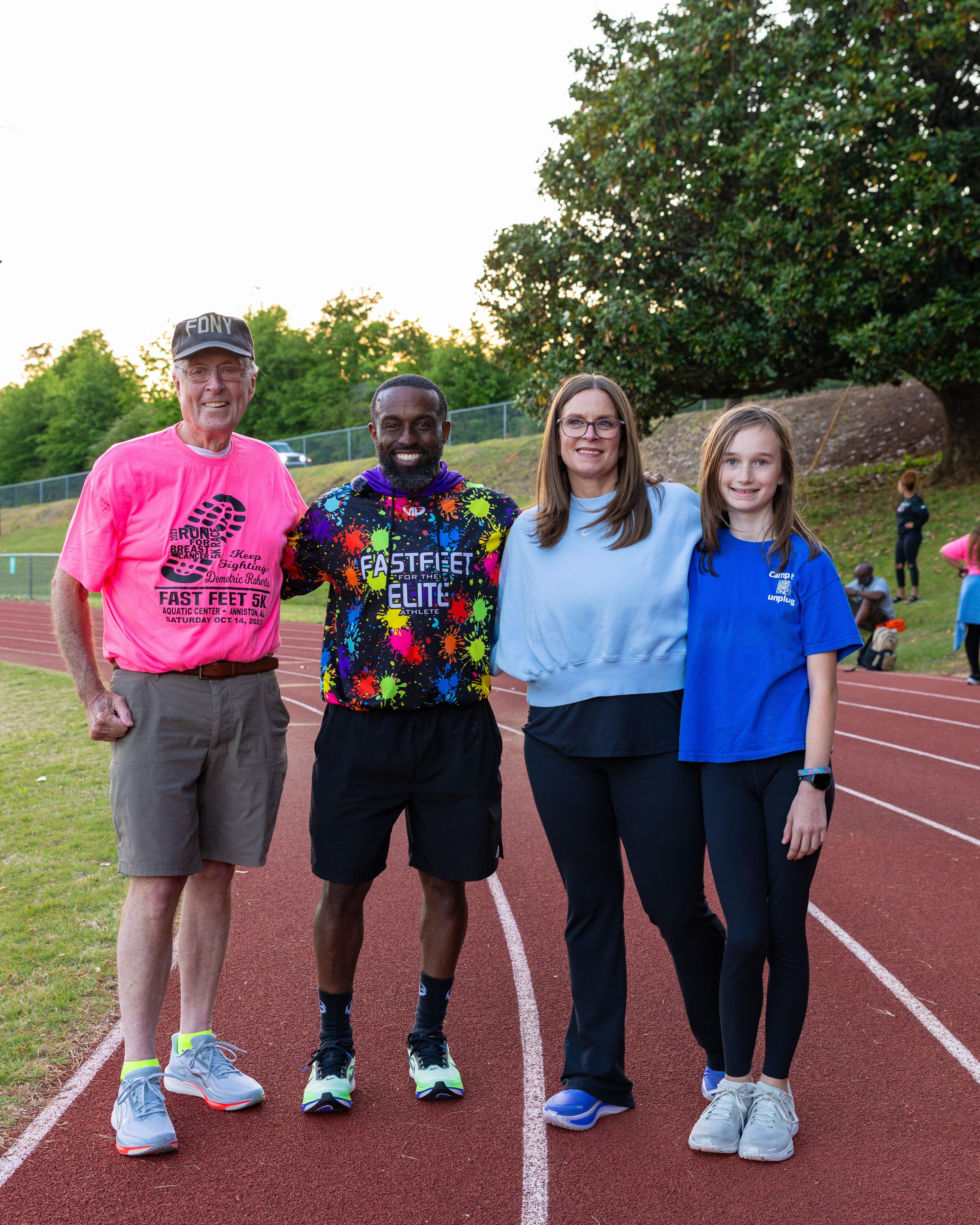 Four people standing side by side on a running track at an outdoor field, with trees and a fence in the background. One person wears a bright pink race shirt, another wears a colorful &ldquo;Fast Feet for the Elite Athlete&rdquo; hoodie, and the others wear casual athletic clothing.