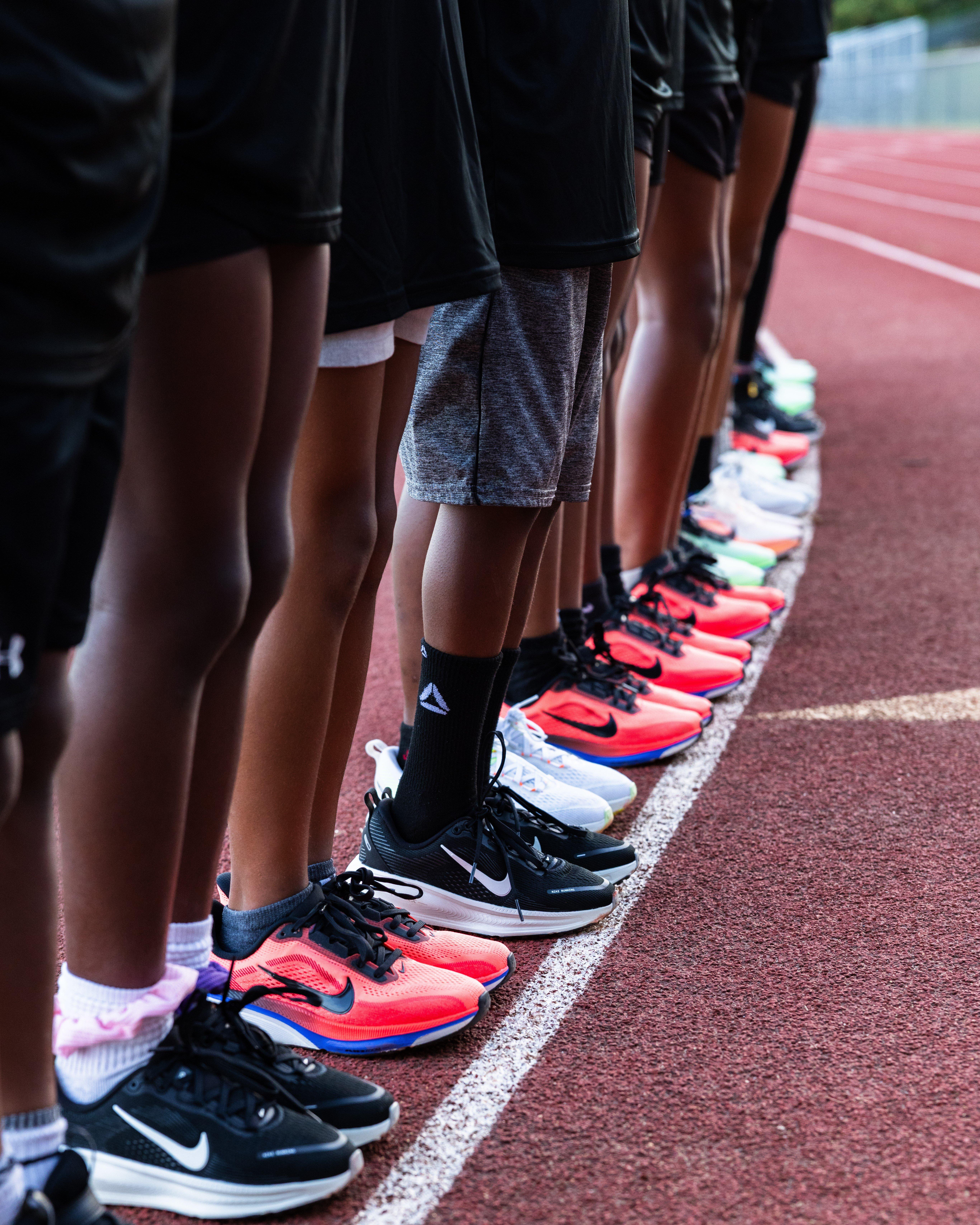 Close-up of a line of young athletes standing shoulder-to-shoulder on a track, showing legs and feet in athletic shorts and various running shoes, including bright and neutral-colored sneakers, positioned along a white starting line.