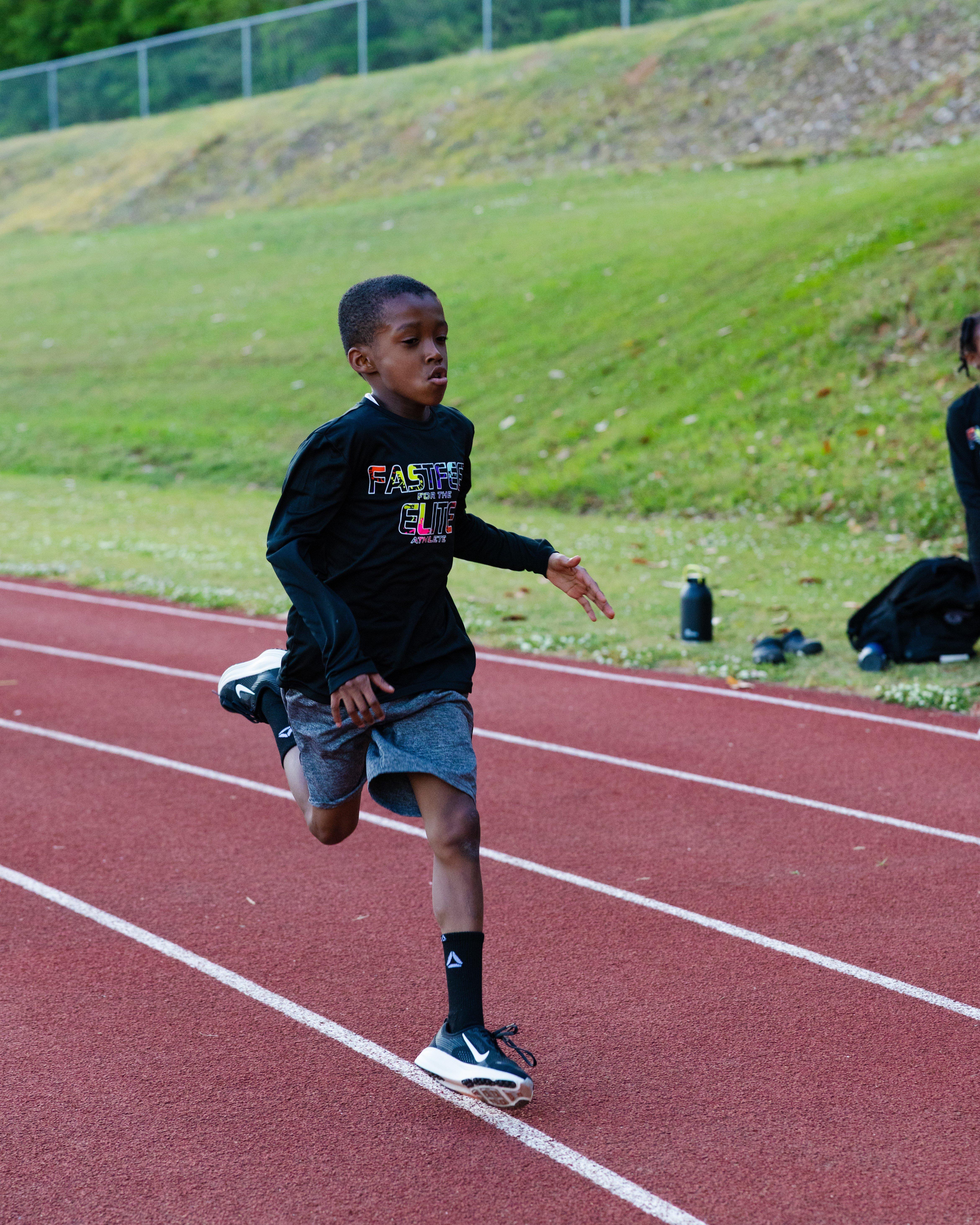 A young athlete sprinting alone on a red track lane, wearing a black long-sleeve &ldquo;Fast Feet for the Elite Athlete&rdquo; shirt, gray shorts, and black running shoes, with grassy hills and a fence in the background.