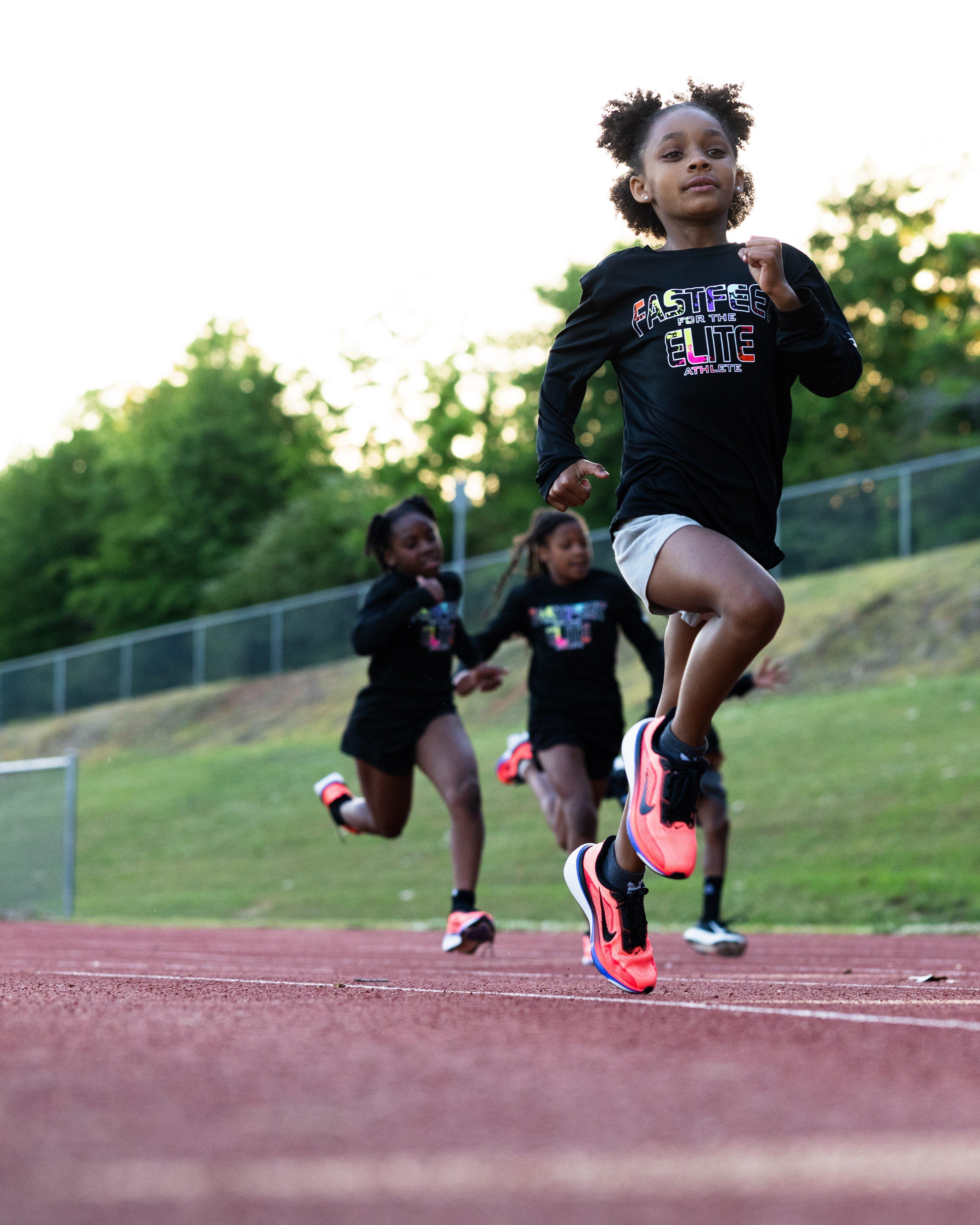 A group of young athletes running on a track, led by one runner in the foreground wearing a black &ldquo;Fast Feet for the Elite Athlete&rdquo; shirt and bright running shoes, while others follow behind on a grassy field beside the track.