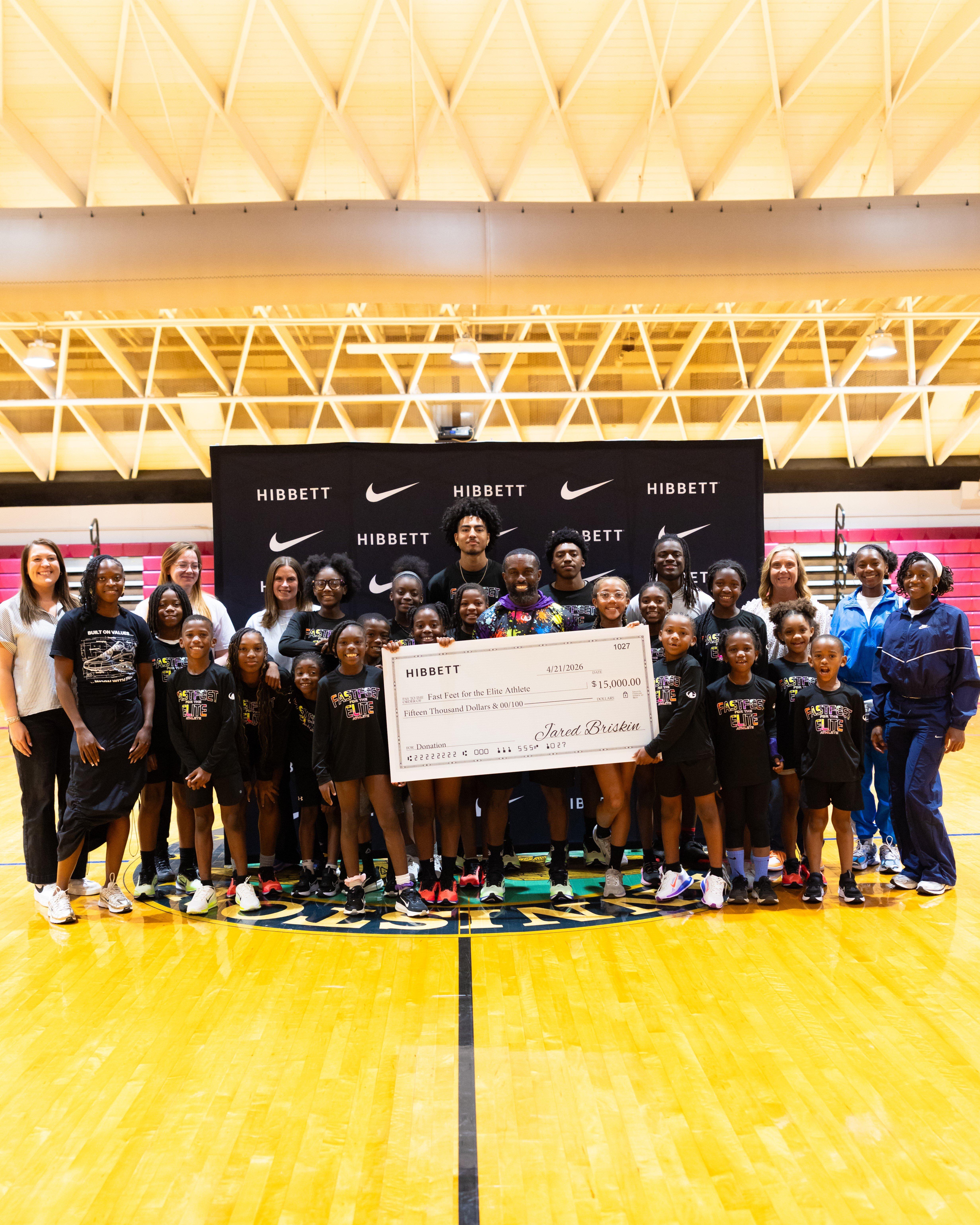 Large group of youth athletes and adults standing on an indoor basketball court in front of a black backdrop with repeating &ldquo;Hibbett&rdquo; and Nike logos, holding an oversized check made out to &ldquo;Fast Feet for the Elite Athlete&rdquo; for $15,000.