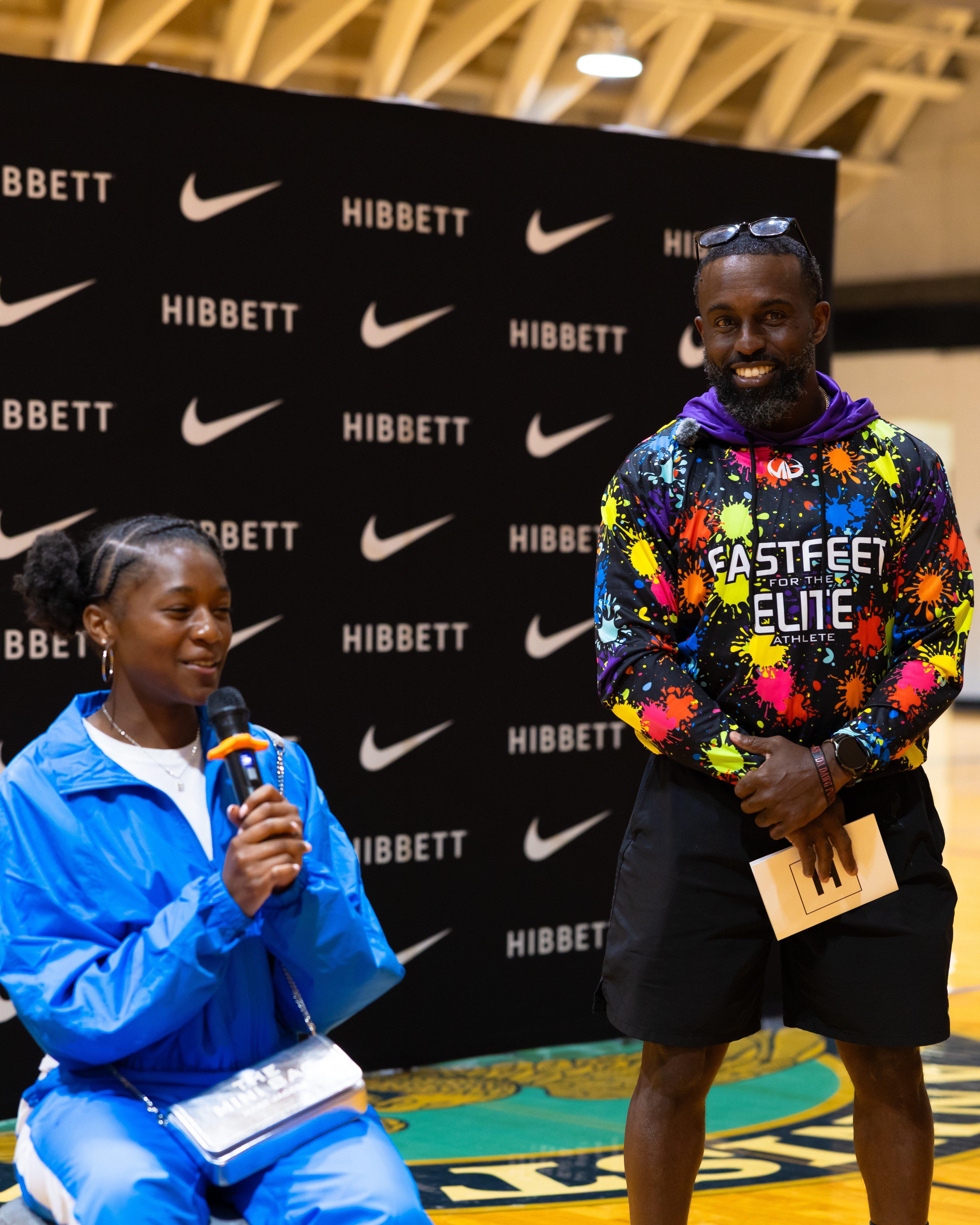 Indoor event scene with one person speaking into a microphone while seated, next to another person standing in a colorful &ldquo;Fast Feet for the Elite Athlete&rdquo; hoodie, both in front of a black backdrop featuring &ldquo;Hibbett&rdquo; and Nike logos.