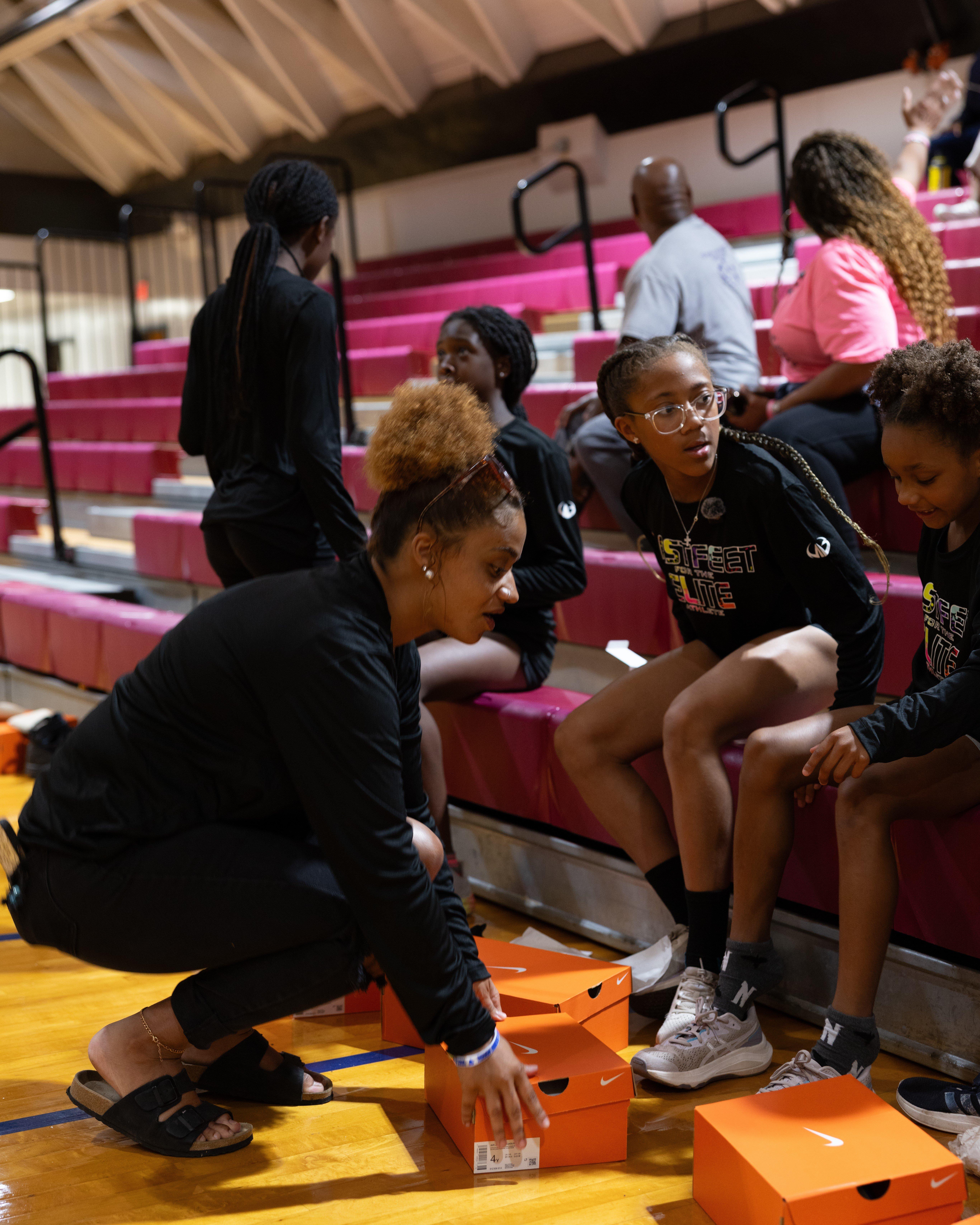 Indoor gym setting where several young athletes sit on bleachers while an adult hands out orange Nike shoe boxes, with more boxes on the floor and bleachers visible in the background.
