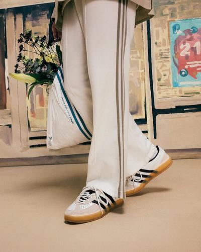 A person is standing, wearing white Adidas pants with brown stripes and Bad Bunny x Adidas Gazelle sneakers. The sneakers are light-colored (possibly white or light grey) with black Adidas stripes and a gum sole. The person is holding a white Adidas tote bag with blue stripes that contains a bouquet of flowers. The background features abstract painted elements.