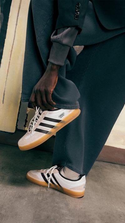 A close-up shot of a person's legs and hands, showcasing Bad Bunny x Adidas Gazelle sneakers. The person is wearing dark pants and holding up one pant leg to better display the white and black sneakers with a gum sole. The other foot is on the ground, also wearing the shoe with a patterned sock. The background is an abstract painted wall.