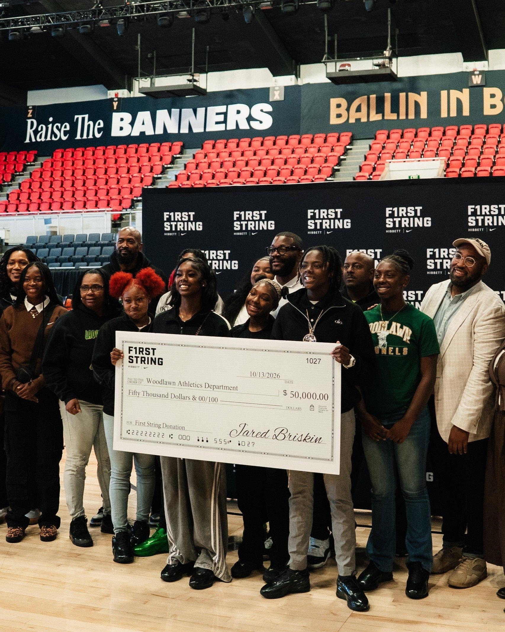 Group holding a $50,000 First String donation check for the Woodlawn Athletics Department on a basketball court.