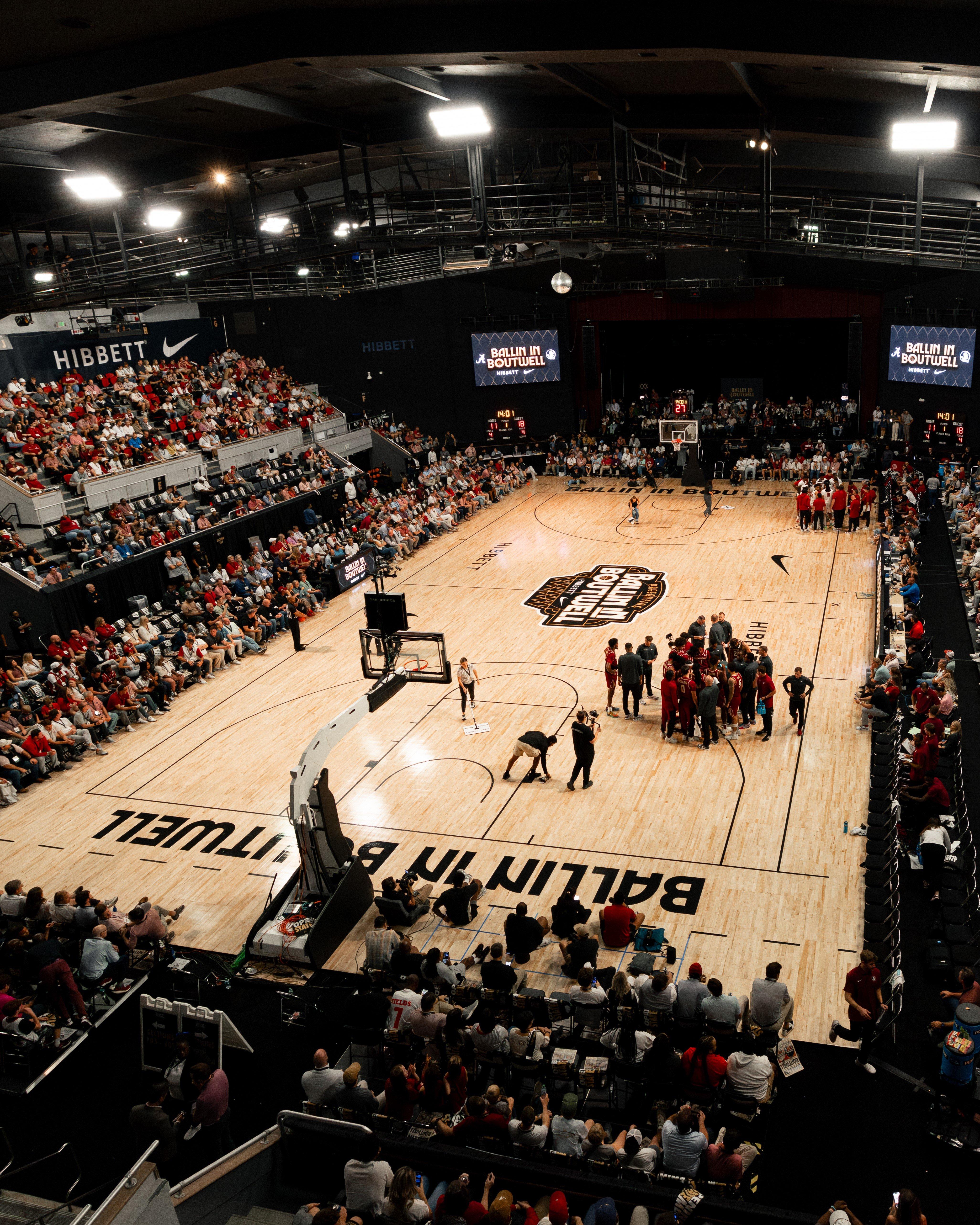 An aerial view of a large, packed basketball arena with spectators filling the stands. The court floor features "BALLIN IN BOUTWELL" text.