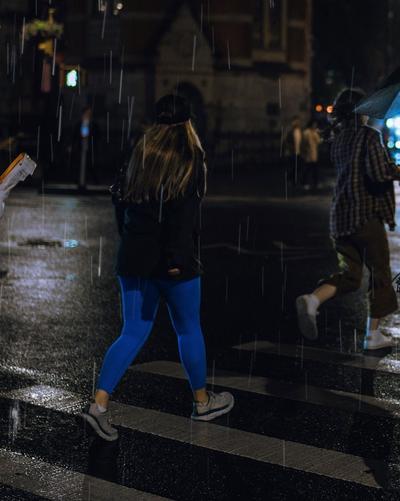 Person wearing bright blue leggings and gray running shoes walking across a wet city street at night in heavy rain as another pedestrian carries an umbrella.