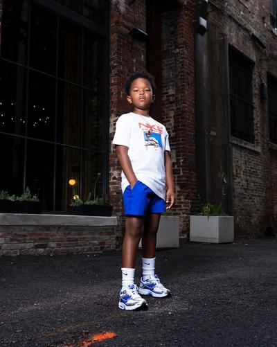 Child wearing a white Nike graphic tee, blue shorts, white crew socks, and blue-and-white Nike sneakers standing on a city street.