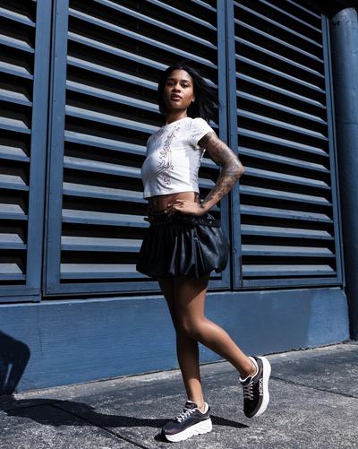 Woman posing outdoors in front of a metal wall wearing a white graphic crop top, black skirt and black athletic sneakers.