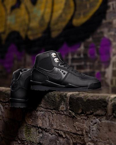 Pair of all-black Nike boots displayed on a stone ledge, showing the textured Swoosh, metal lace hooks and rugged traction outsole.