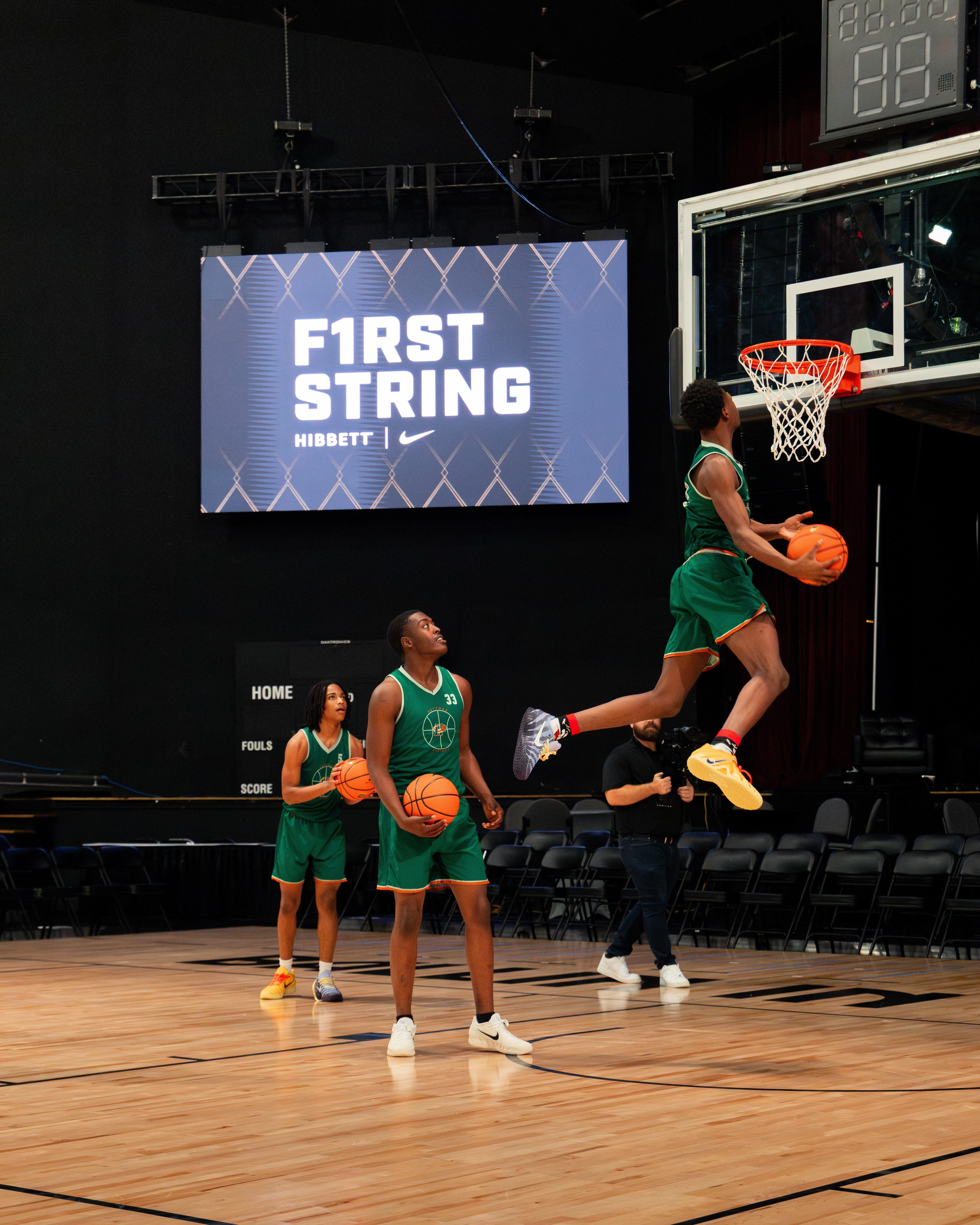 A male basketball player in a green jersey and shorts is mid-air executing a dunk, with two teammates looking on. A large screen in the background displays the F1RST STRING HIBBETT logo.
