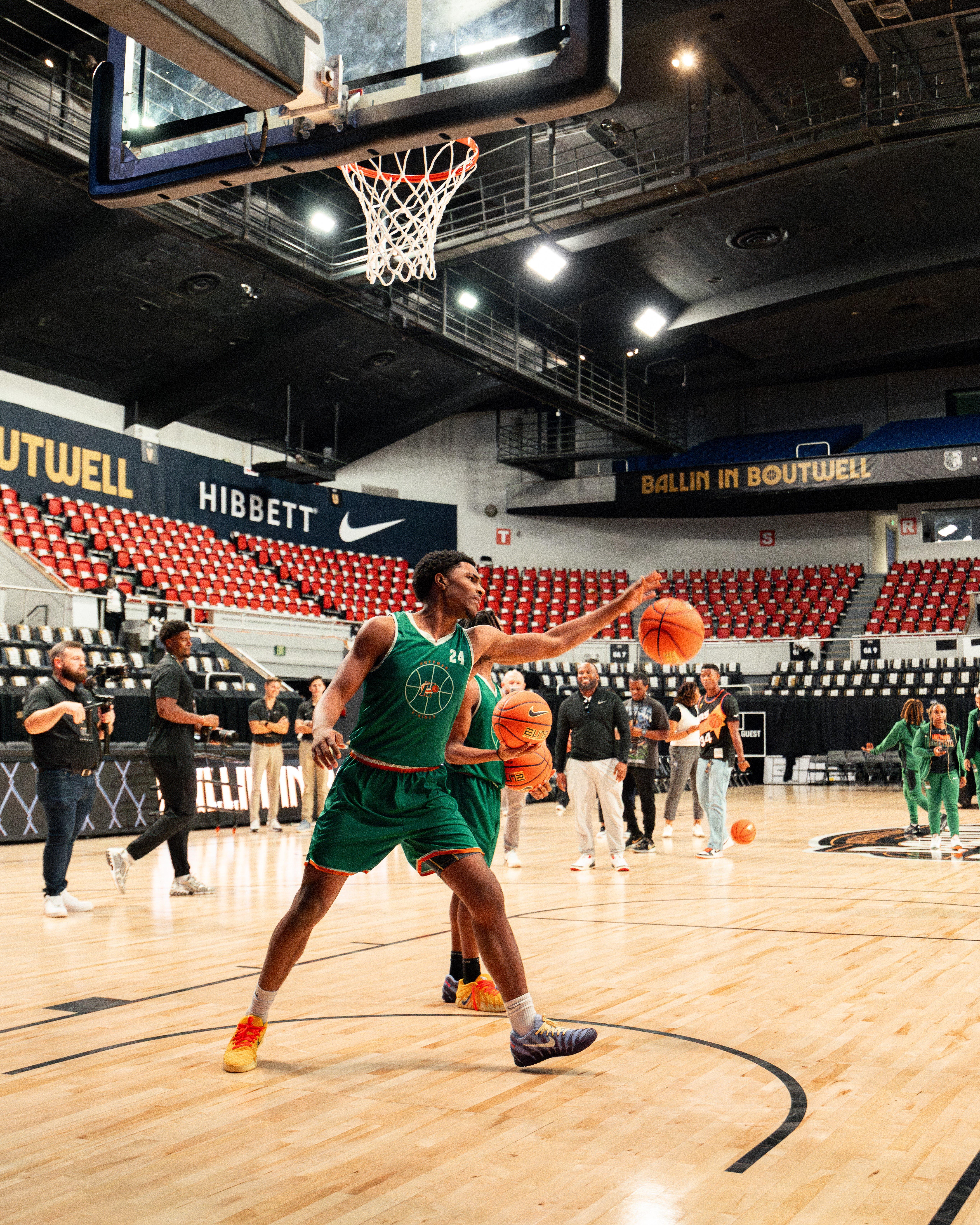 A male basketball player in a green uniform is dribbling a ball on a wooden court in an arena, with a crowd of people and camera crew visible in the background.