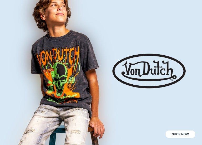 A young boy wearing a faded black Von Dutch graphic tee with a green skull and orange accents, paired with distressed jeans. The Von Dutch logo is featured prominently on the right, with a SHOP NOW button at the bottom.