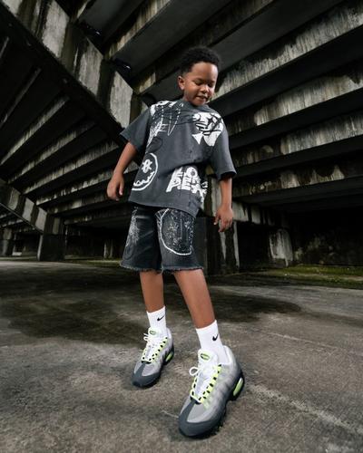 A young boy stands in an urban concrete setting wearing a graphic charcoal t-shirt, matching denim shorts, white Nike crew socks, and grey Nike Air Max 95 sneakers with neon green accents.