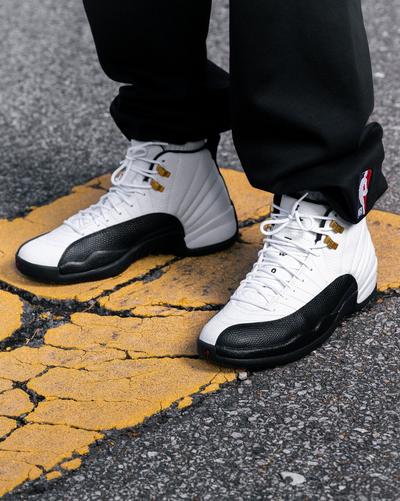 White and black Air Jordan 12 Retro sneakers with gold lace hardware worn over cracked yellow pavement.