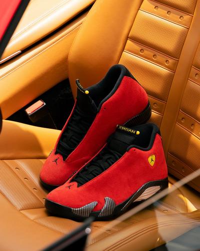 Pair of red suede Air Jordan sneakers with black laces, yellow branding and carbon-fiber accents displayed on a tan leather car seat.