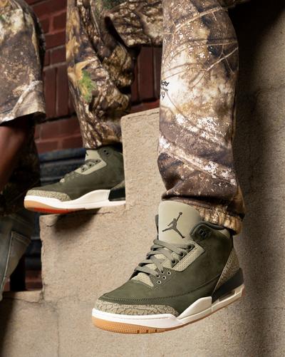 Olive green Air Jordan 3 Retro sneakers with tonal elephant-print accents styled with camouflage apparel on outdoor steps.