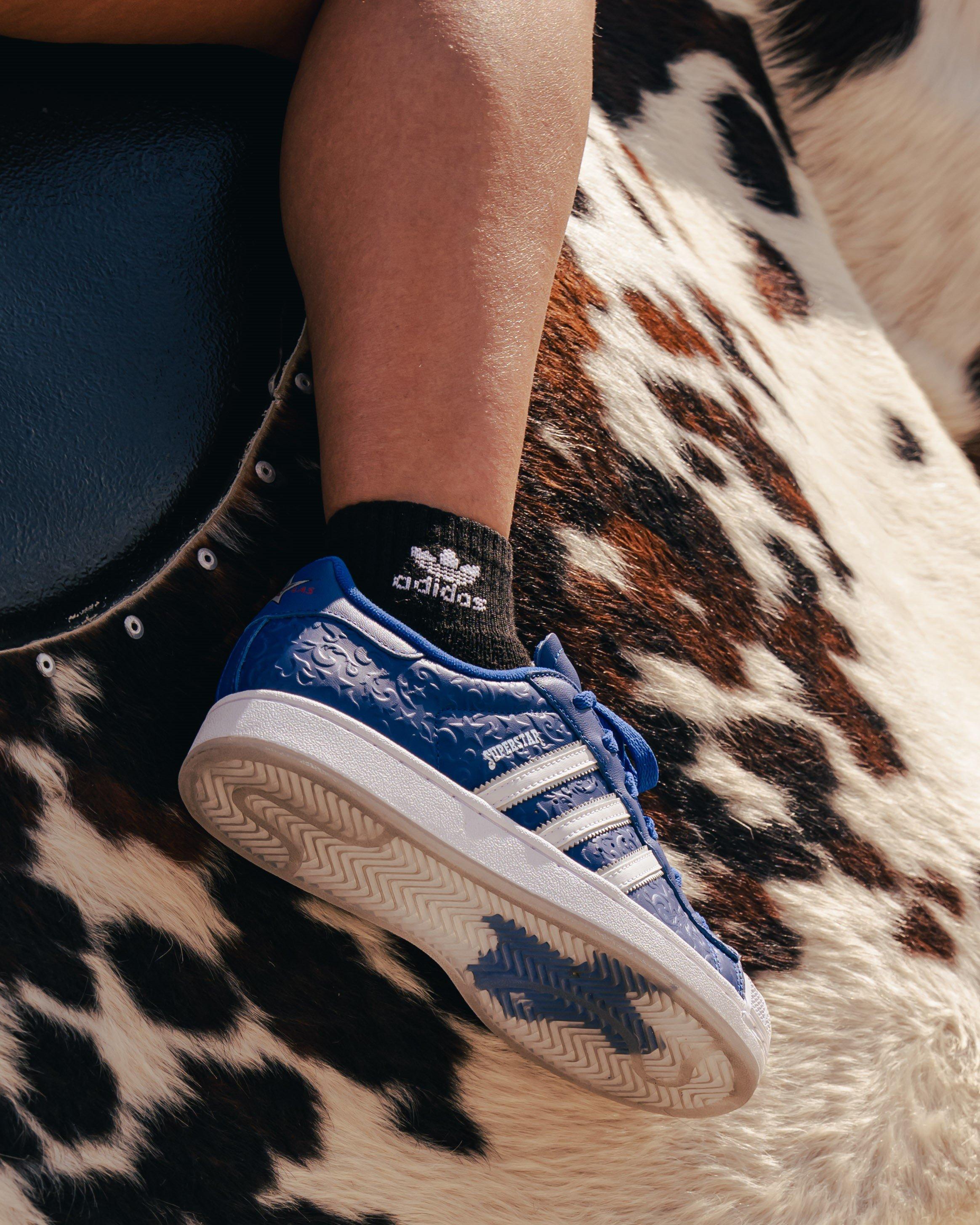 A close-up of a person's leg and foot resting on a mechanical bull covered in faux cowhide. The person is wearing a gray sock with an Adidas logo and a blue Adidas Superstar sneaker with a white shell toe and stripes.