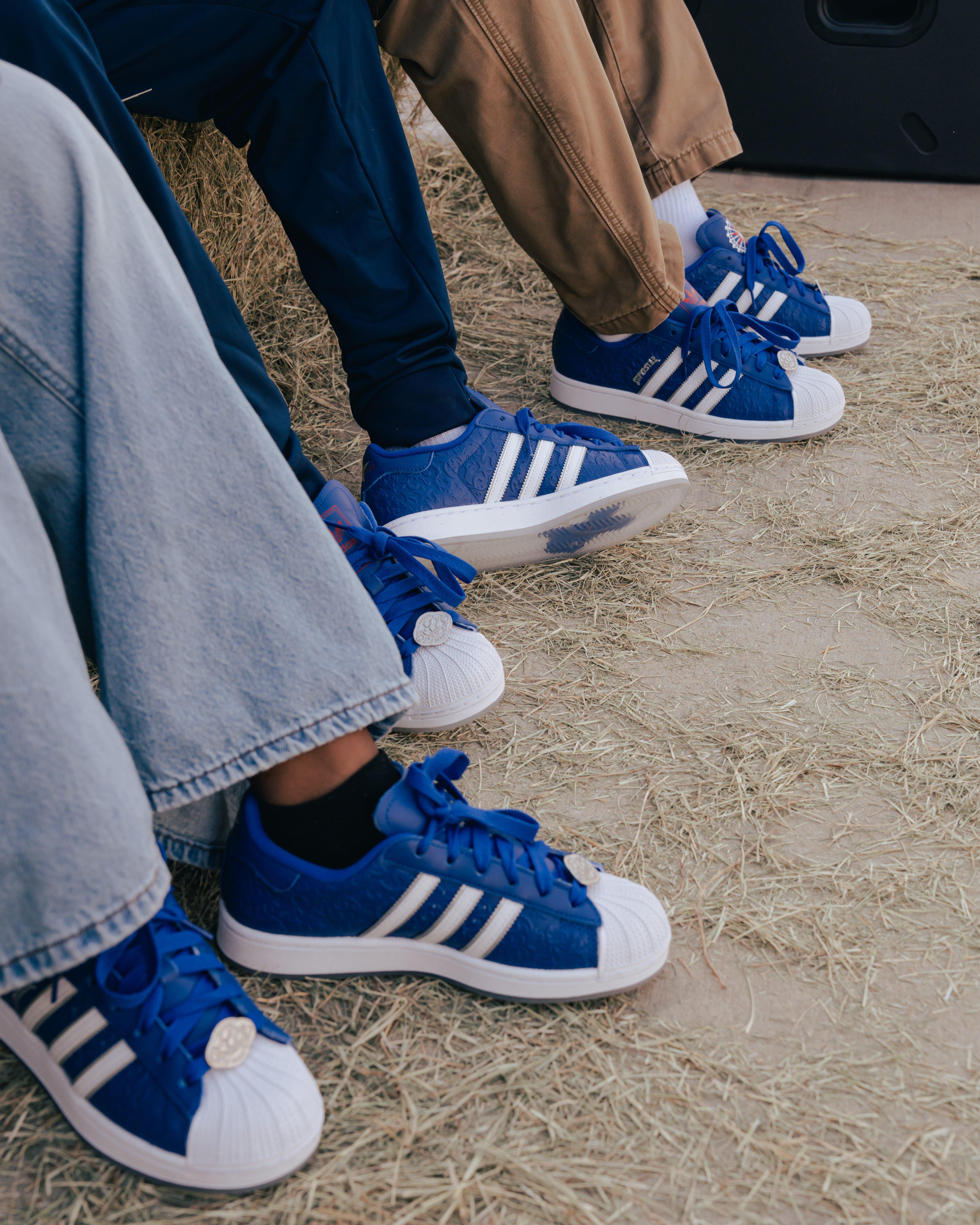 A close-up shot of three people's feet sitting on a floor covered in hay. Each person is wearing a blue Adidas Superstar sneaker with white stripes. Two of the pairs have silver Texas-shaped charms on the laces