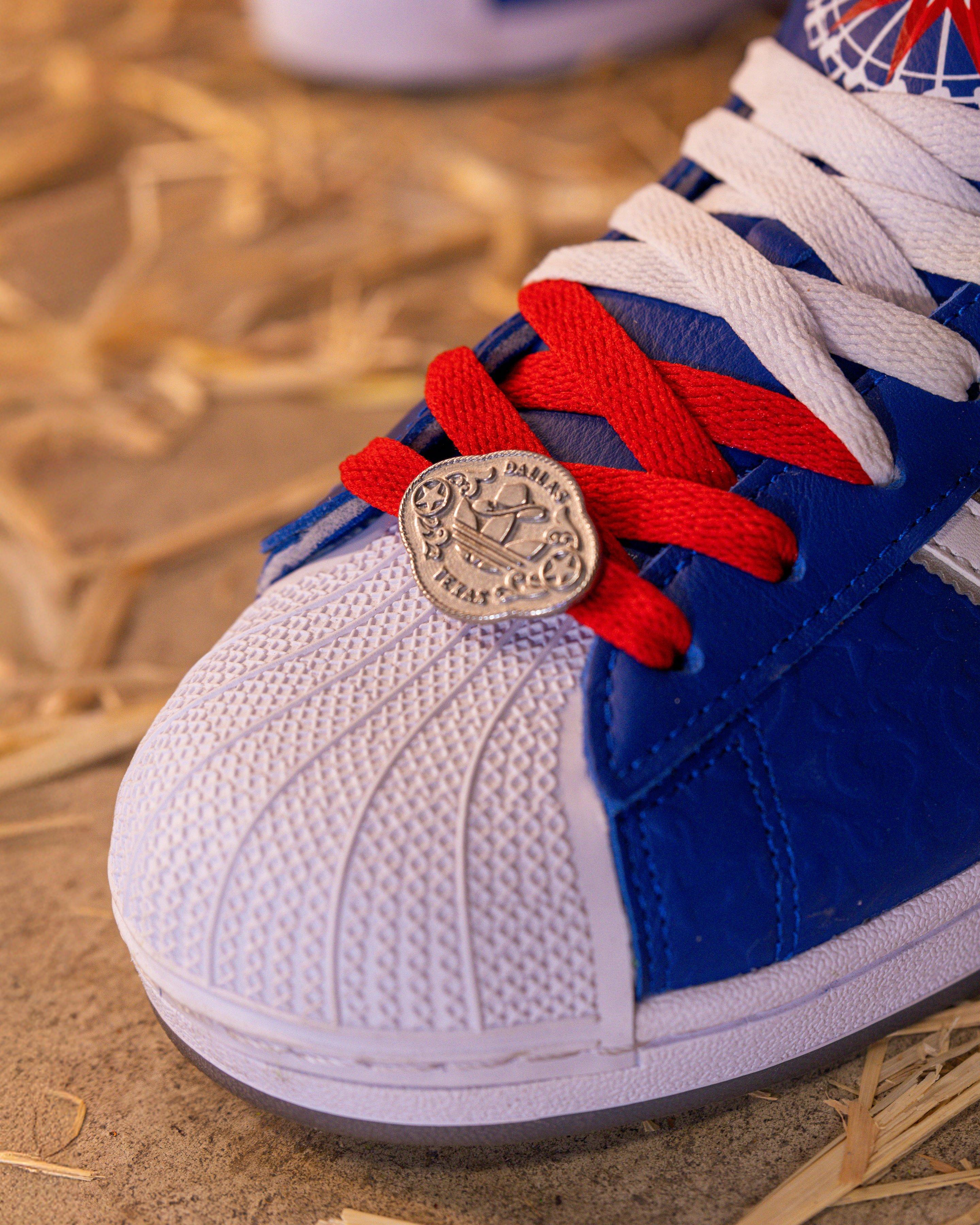 A close-up shot of the toe of a blue and white Adidas Superstar shoe. The laces are tied with a red lace and a white lace, and a silver coin charm with "DALLAS TEXAS" is attached to them. The white shell toe has a ribbed pattern.