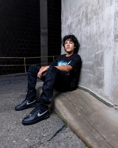 Person wearing a graphic T-shirt, black jeans and black Nike sneakers while sitting against a concrete wall in an urban setting.