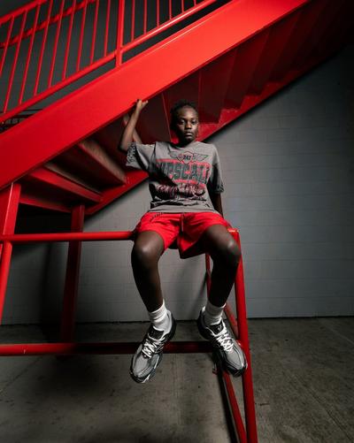 Kid wearing a graphic T-shirt, red shorts and silver sneakers while sitting beneath a bright red staircase.