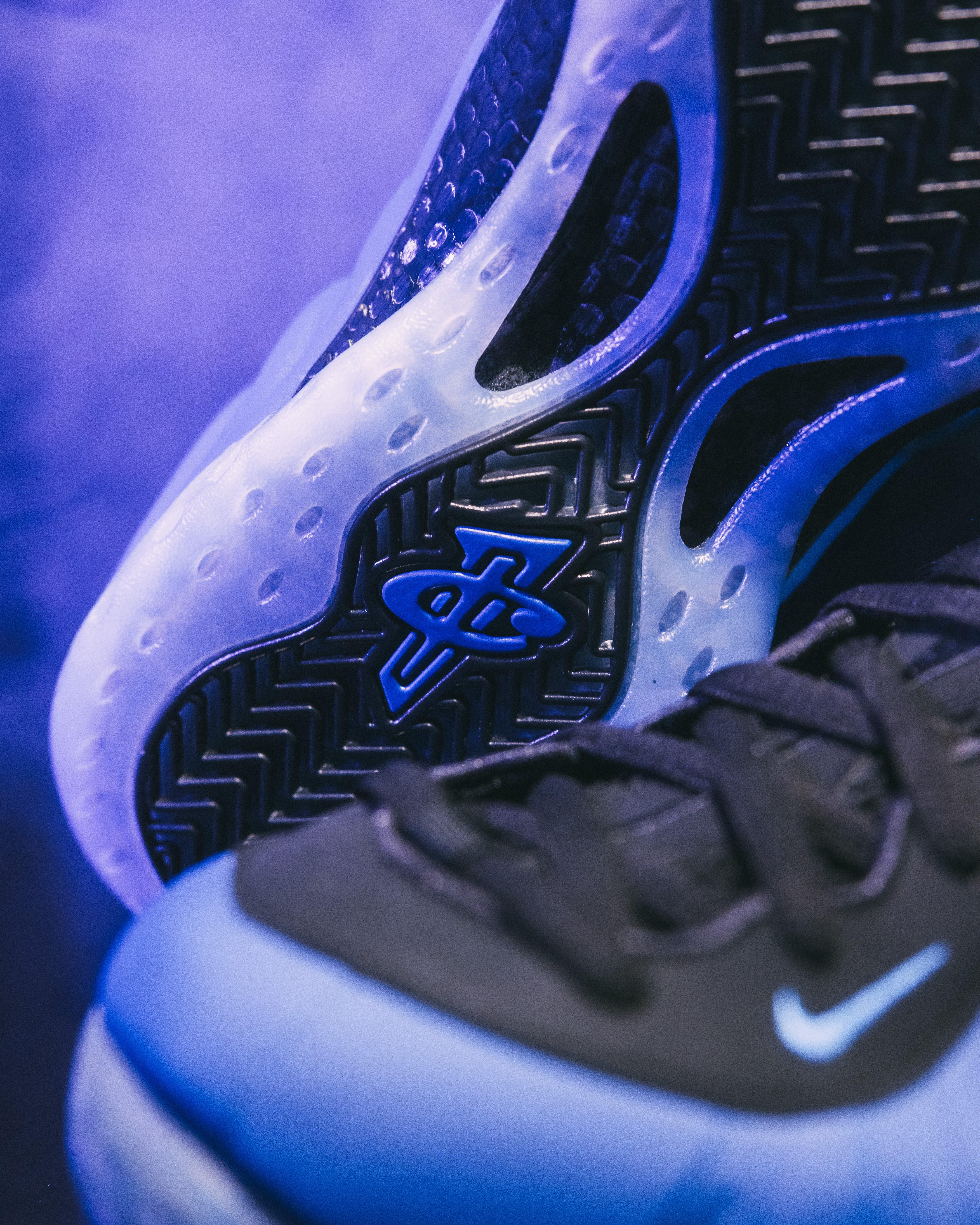 Close-up view of a Nike Foamposite Deep Royal sneaker highlighting the sole and part of the upper. The sole features a distinctive pattern with a blue Nike 'Penny' logo, while the upper is black with blue accents and a small Nike swoosh. The background has a bluish hue, creating a cool-toned atmosphere.