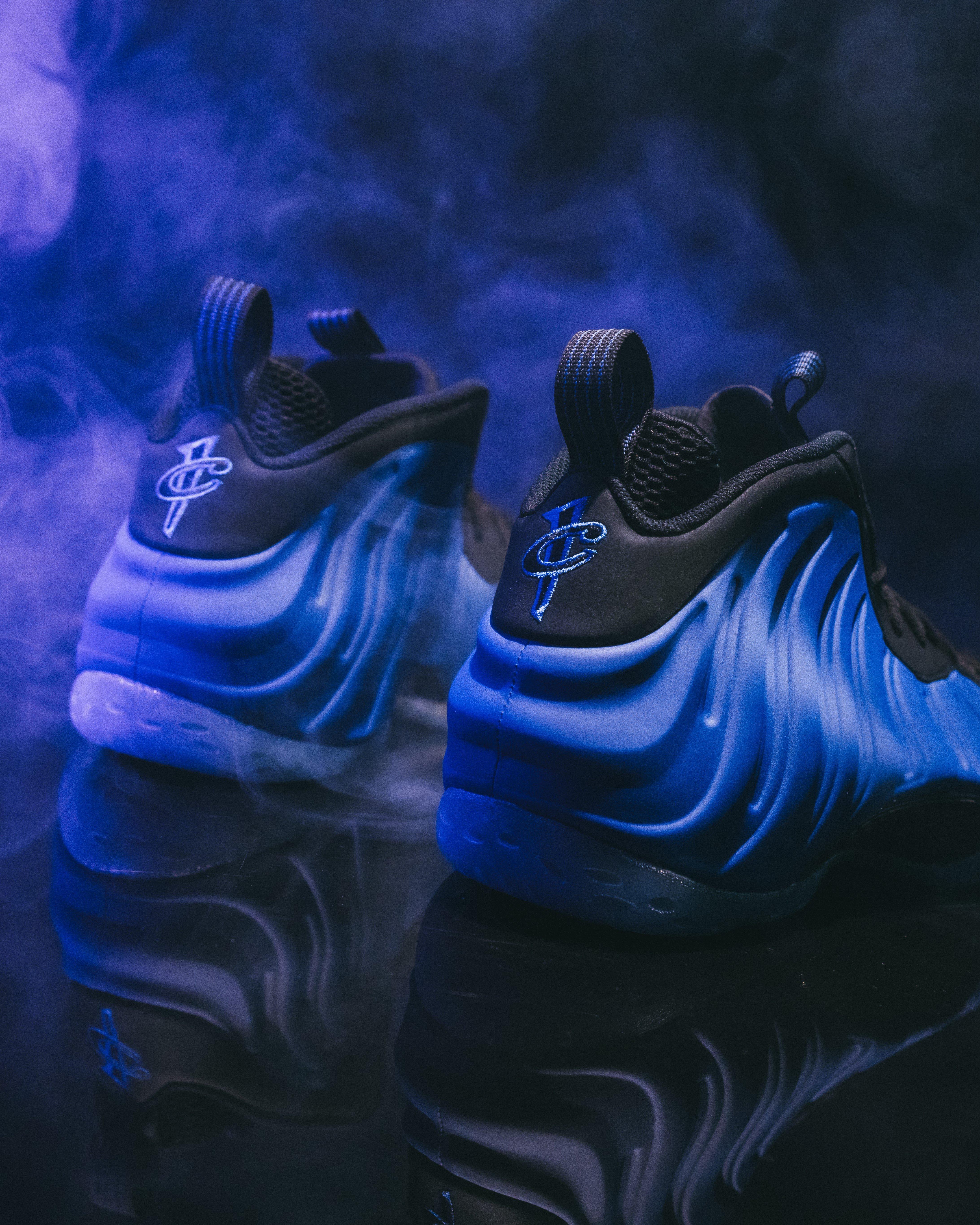 A pair of blue and black Nike Foamposite Deep Royal sneakers displayed on a reflective surface. The shoes feature a wavy side pattern and a logo on the heel. Blue smoke or mist surrounds them against a dark background, creating a dramatic visual effect.