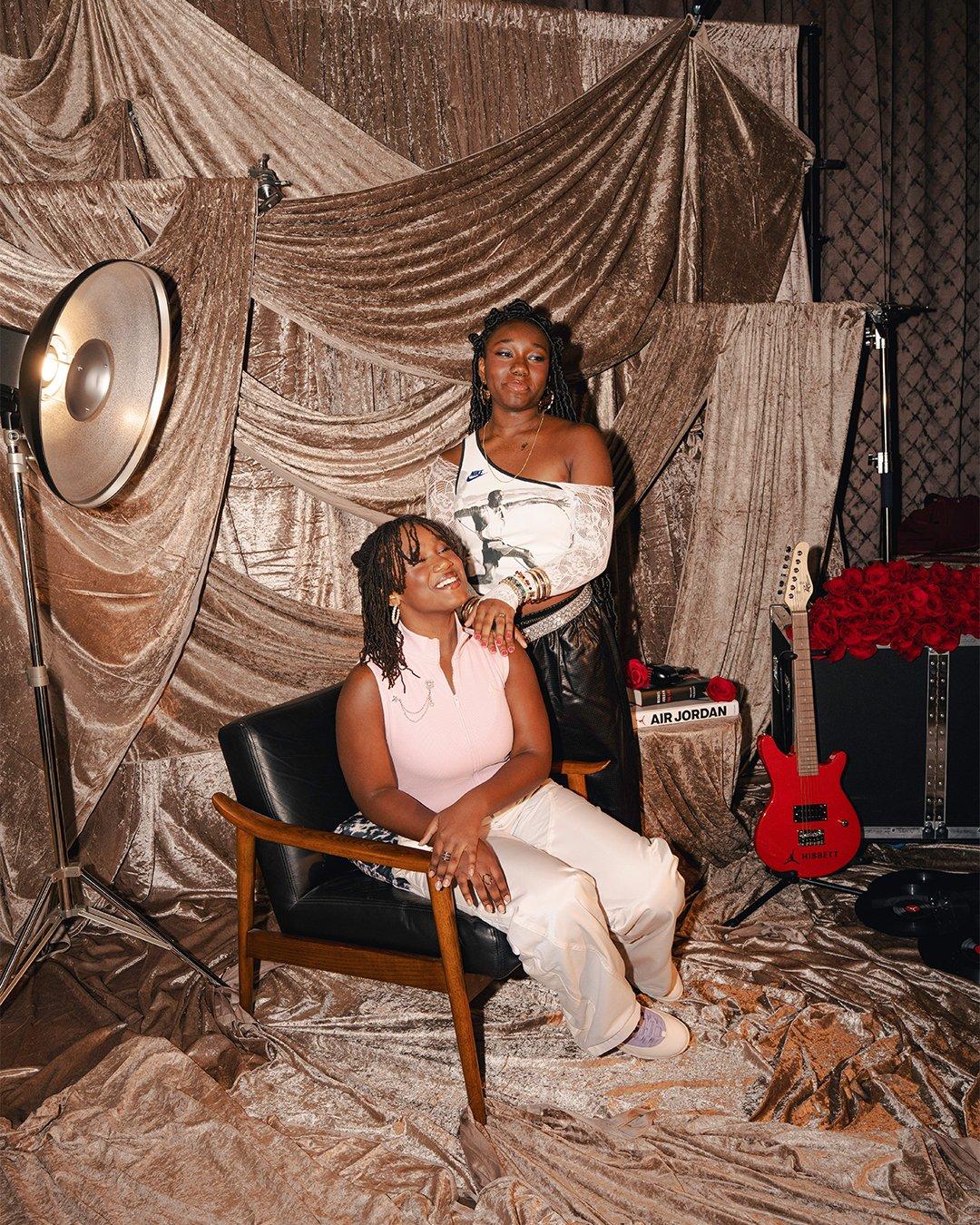Two women pose against a draped, champagne-colored velvet backdrop. One sits in a black leather chair wearing a pink zip-up top and white pants, while the other stands behind her with a hand on her shoulder, wearing a white Nike top and black leather trousers. A red electric guitar and a bouquet of red roses sit to the side.
