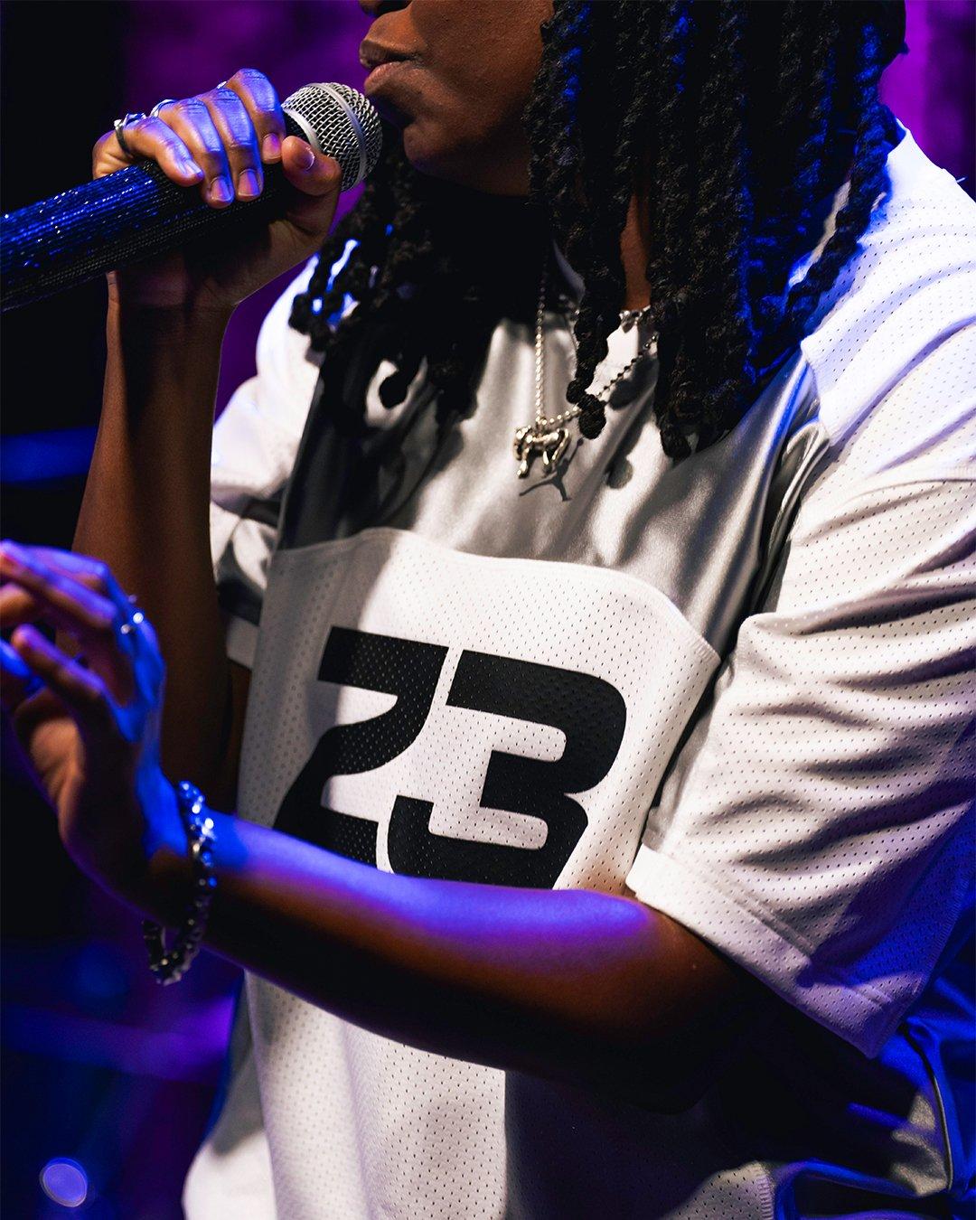 A close-up shot of a person holding a glittery microphone to their mouth. They are wearing a white and grey Jordan Brand jersey with the number 23 and a silver Jumpman pendant necklace. The scene is illuminated by moody purple and blue stage lighting.