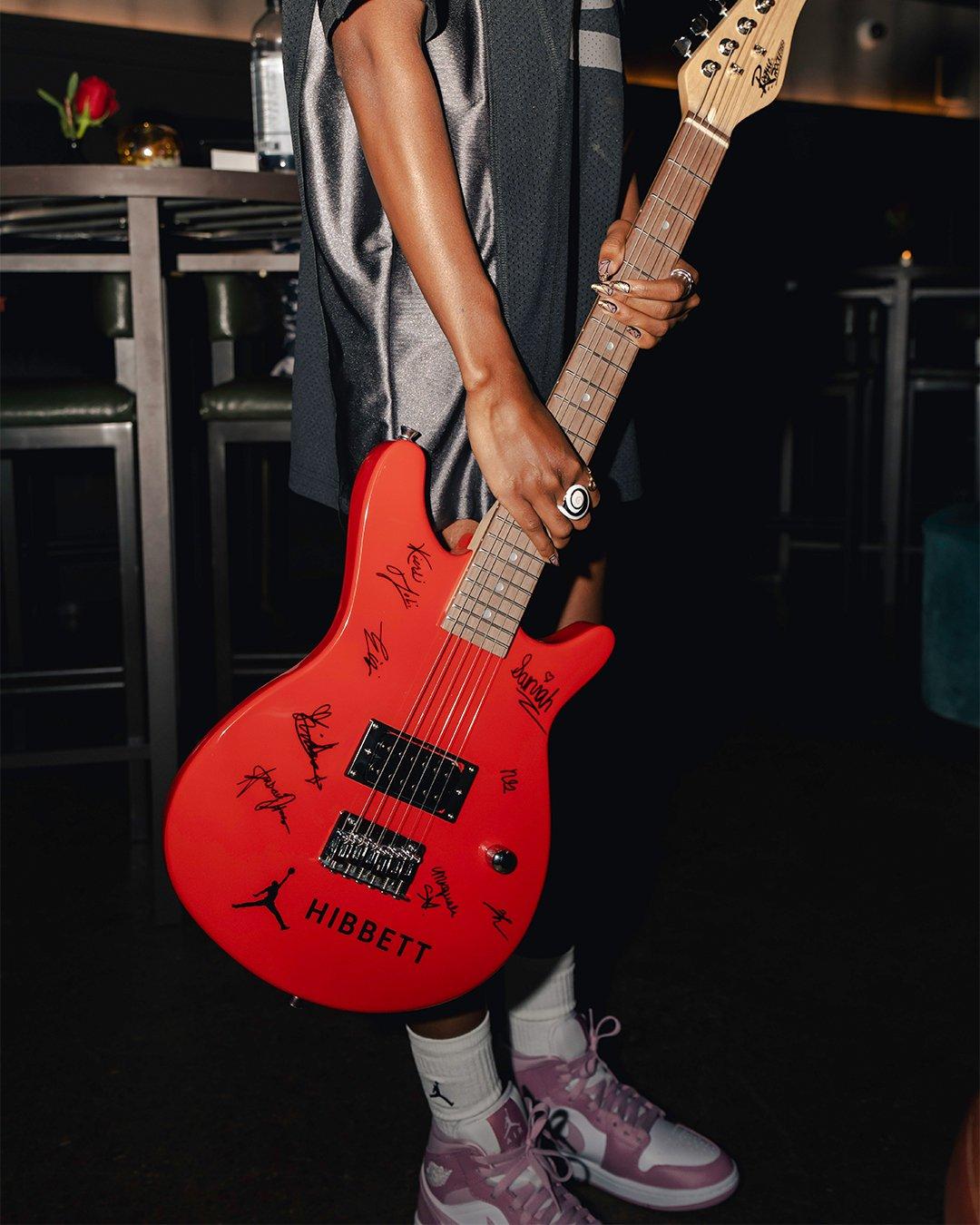 A close-up shot of a person holding a bright red electric guitar covered in black signatures. The guitar features a Jumpman logo and "HIBBETT" branding. The person is wearing a dark satin jersey, white Jordan socks, and pink and white sneakers.