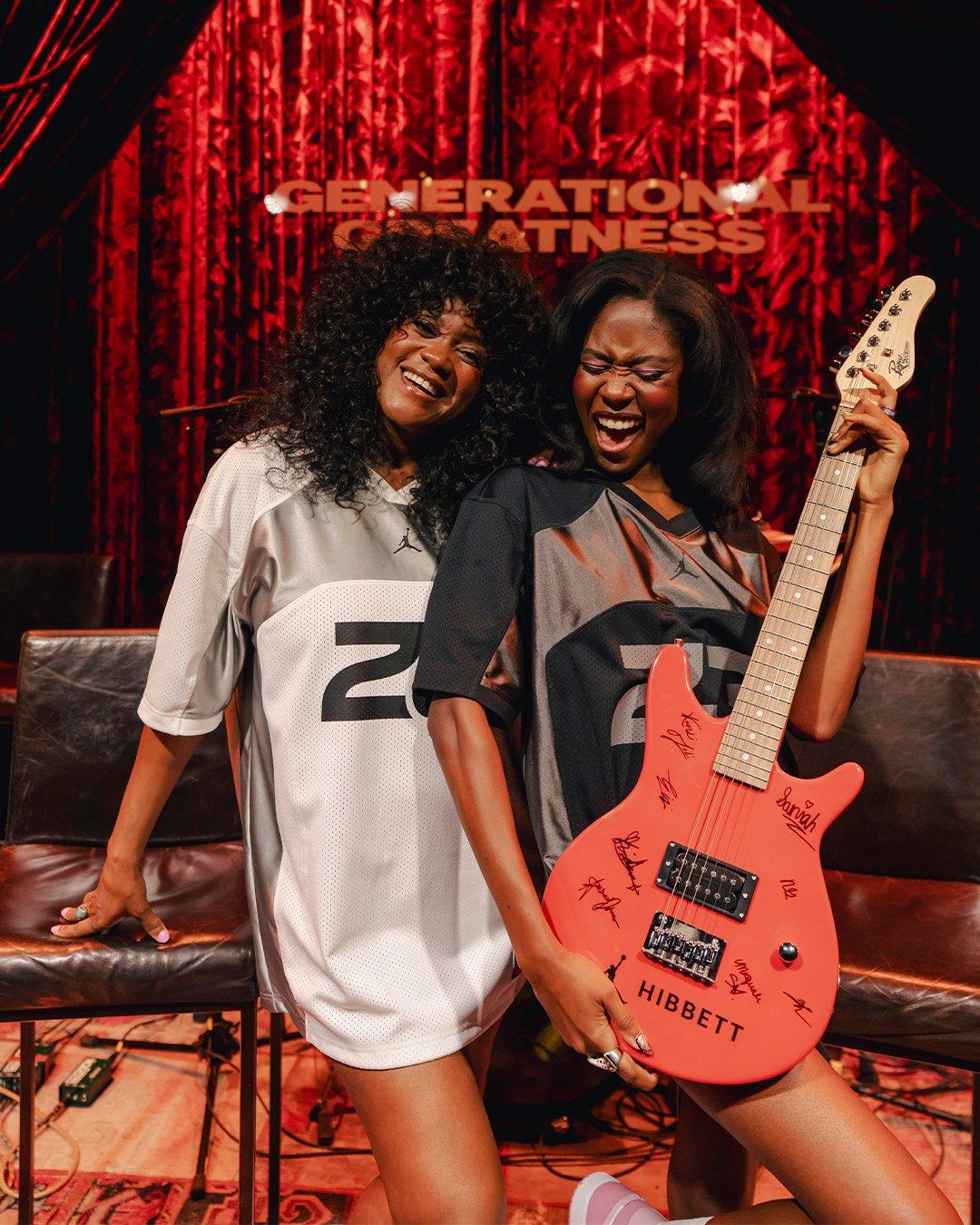 Two  women pose joyfully in front of a red "Generational Greatness" stage backdrop. They are wearing oversized Jordan brand jerseys.