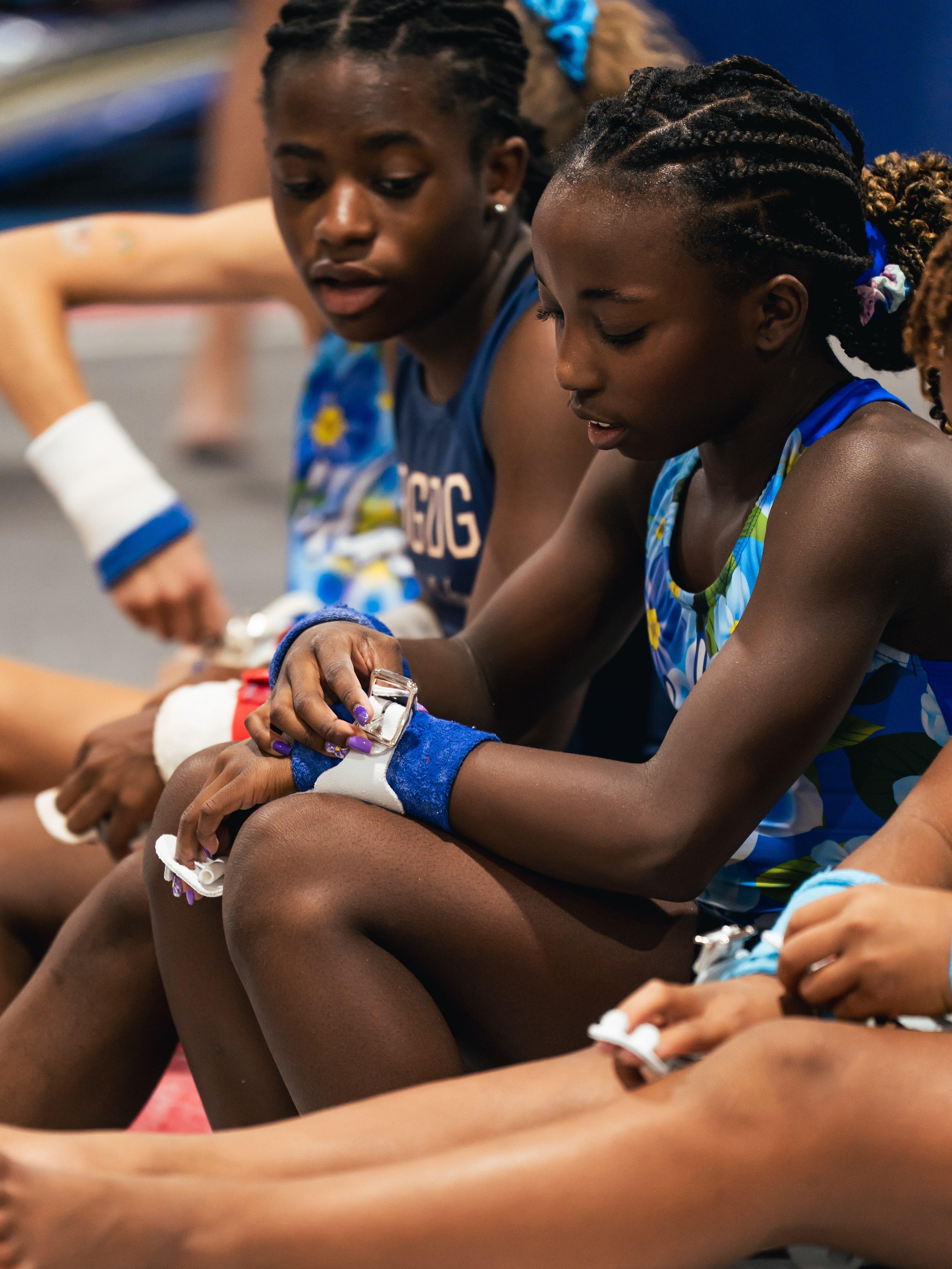 Two young female gymnasts sit side-by-side. The girl on the right, with braided hair, is focused on adjusting her wrist guards. The girl on the left looks toward her. Both are wearing blue and white leotards.