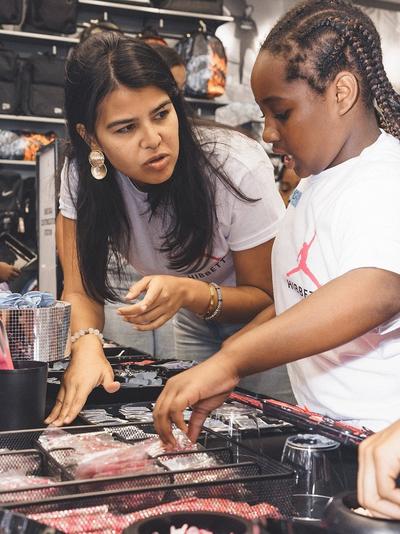 A woman with long dark hair and large gold earrings is helping a young girl select items from a tray to customize a backpack. Both are wearing white t-shirts.