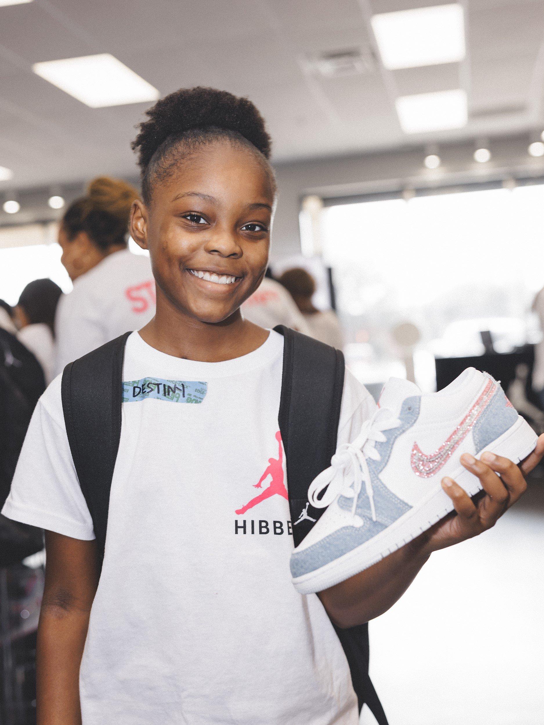 A young girl with a name tag that says "DESTINY" smiles at the camera while holding a white and light blue Jordan sneaker with a sparkling pink swoosh. She is wearing a white t-shirt with the Jordan and Hibbett logos and a black backpack.