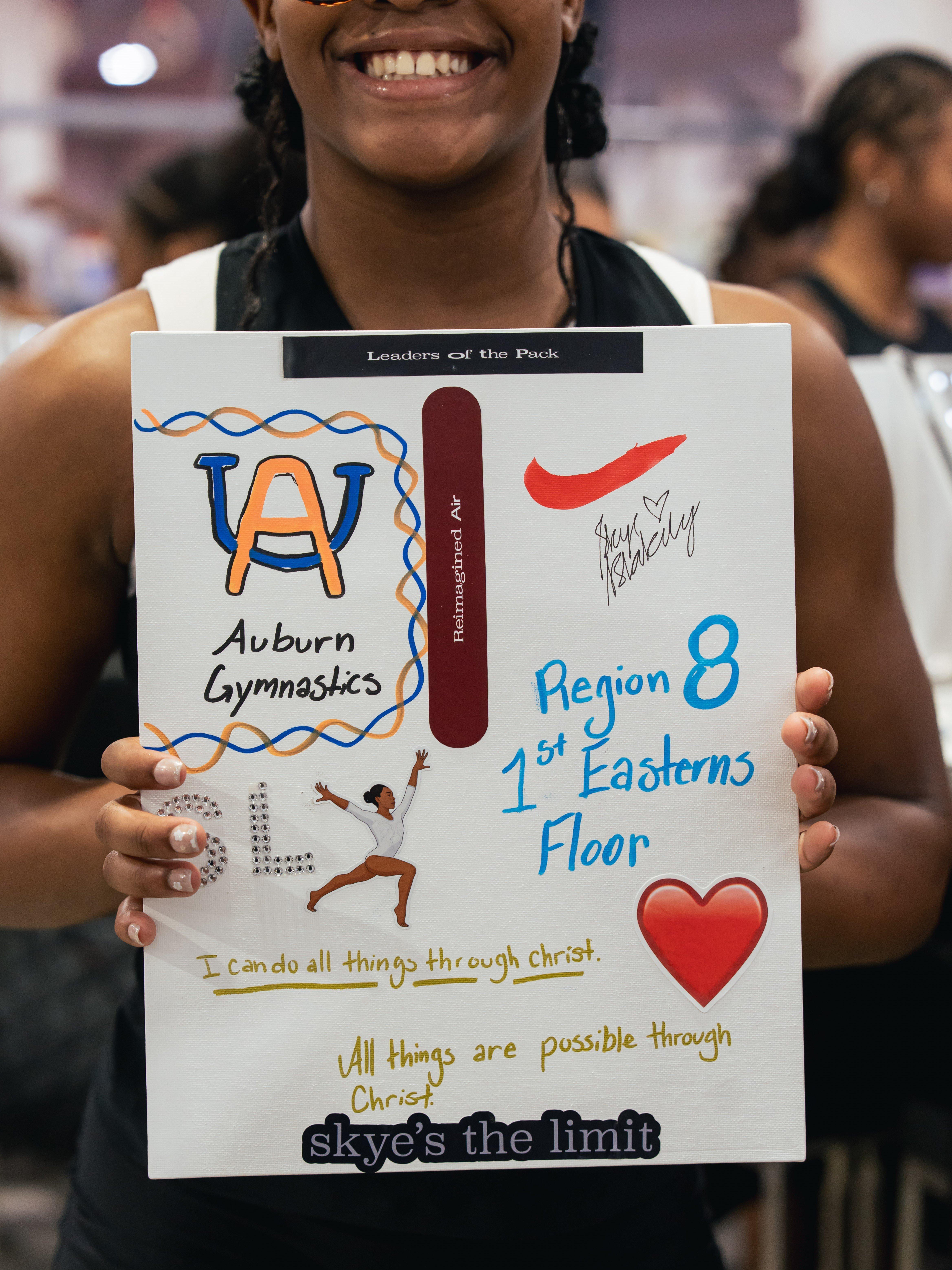 A close-up shot of a young girl's hands holding up a vision board. The board has various handwritten and drawn elements, including a Nike swoosh, the phrase "Auburn Gymnastics," and the quote "I can do all things through christ."