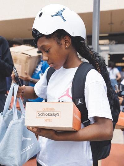 A young girl with a white helmet featuring a blue Jordan logo and a backpack holds a box from Schlotzsky's and a tote bag, while looking at her phone.