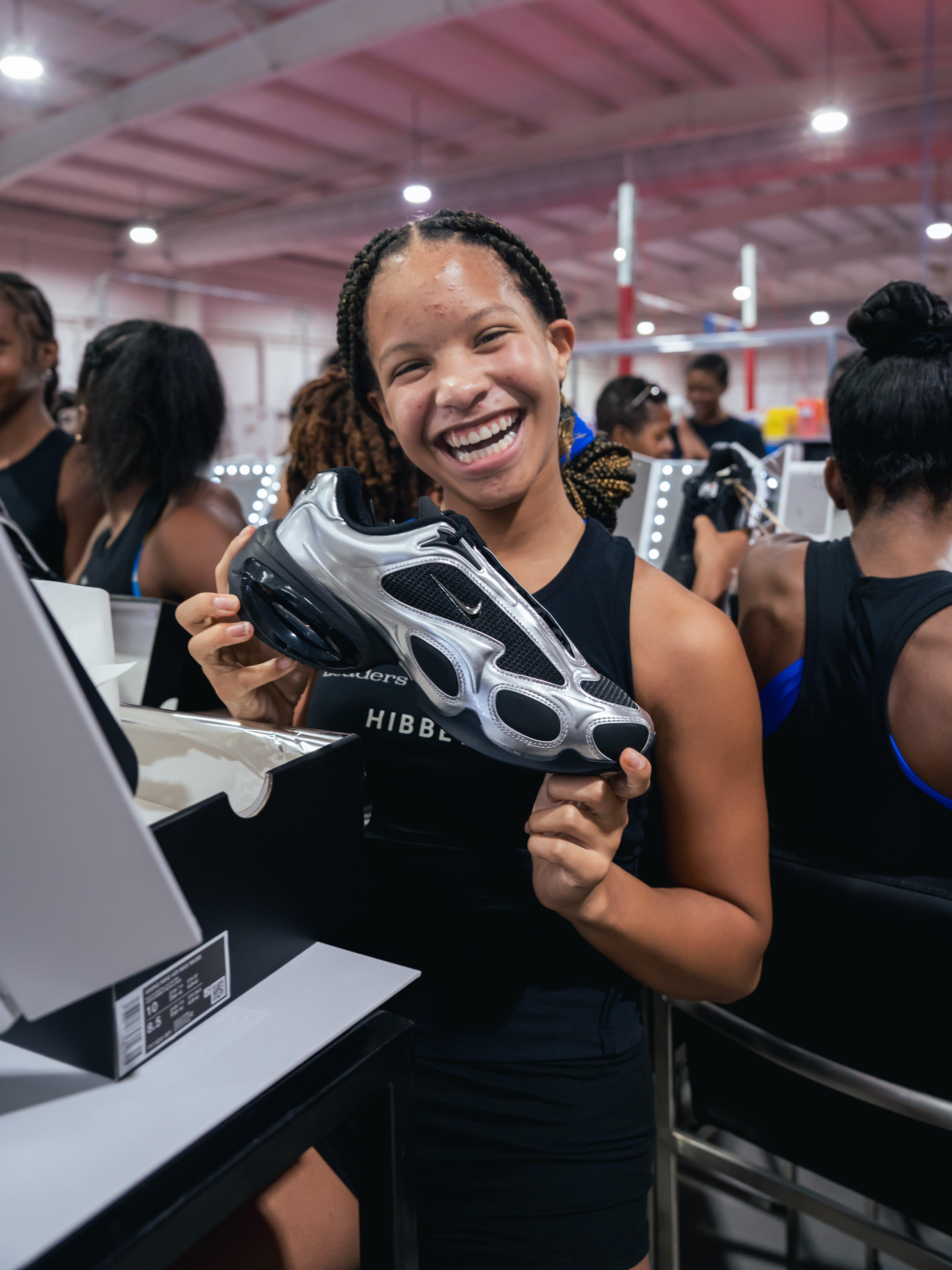 A smiling teenage girl with braided hair holds up a black and silver Nike Air Max Muse shoe. She is wearing a black tank top with a logo.