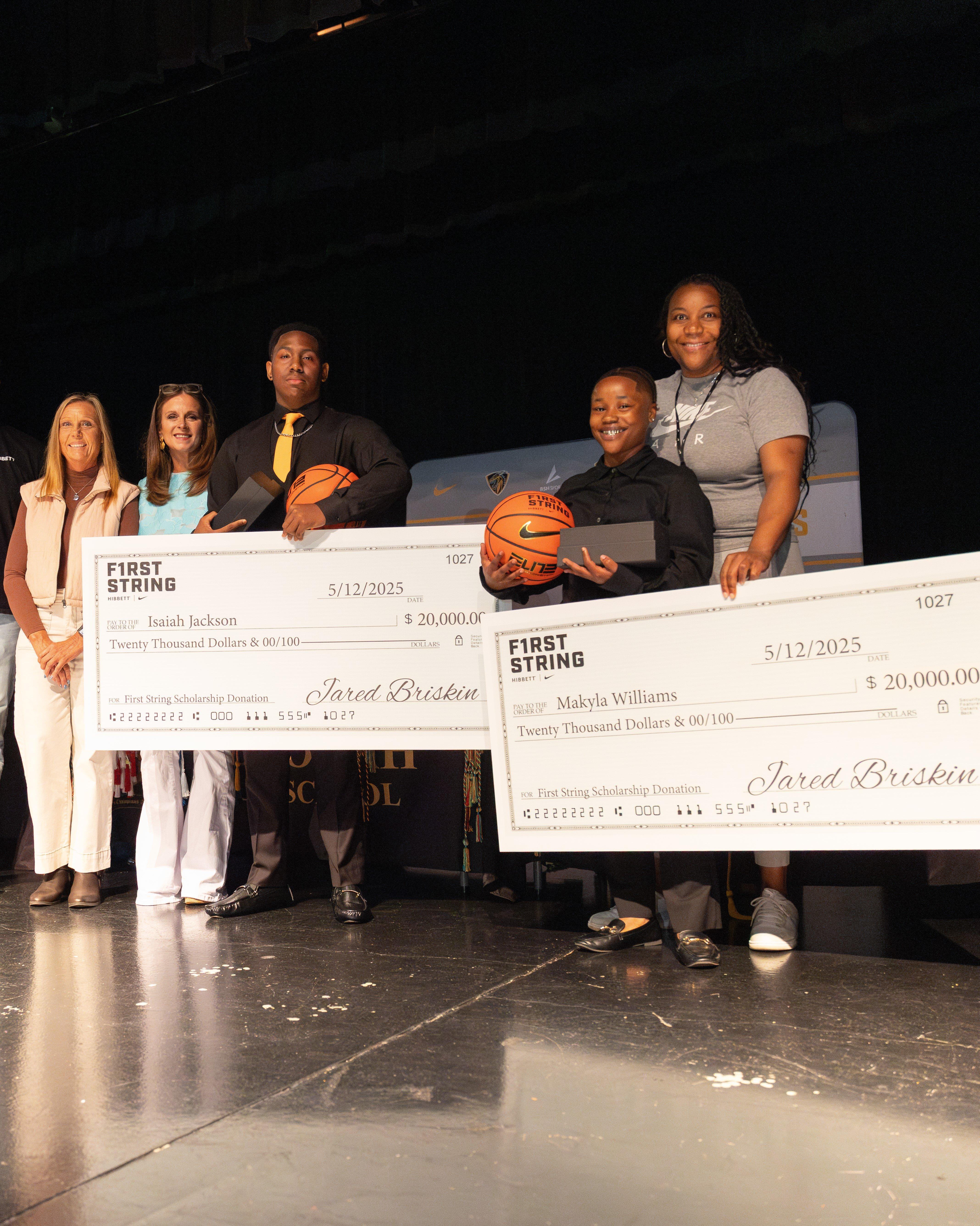 Two young students, dressed in button-up shirts and ties, stand on a stage holding oversized scholarship checks from 'First String Scholarship Donation'. Each student also holds a basketball. Behind them, a woman in a grey t-shirt looks on. The checks are dated '5/12/2025' and are for 'Twenty Thousand Dollars'.