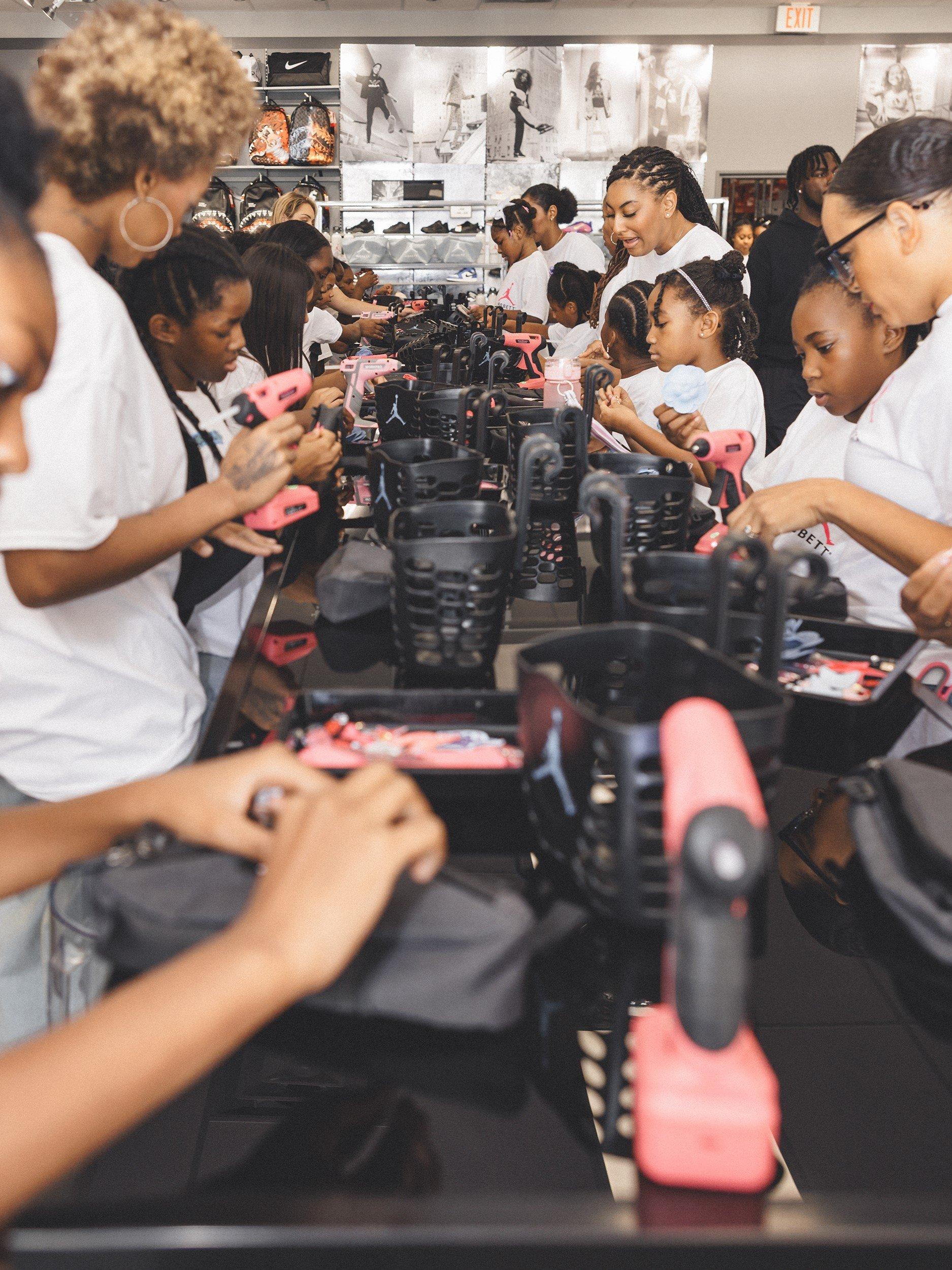  A group of adults and children in a store are sitting at a long table, customizing backpacks with small drills. They are all wearing white t-shirts with the Jordan and Hibbett logos.