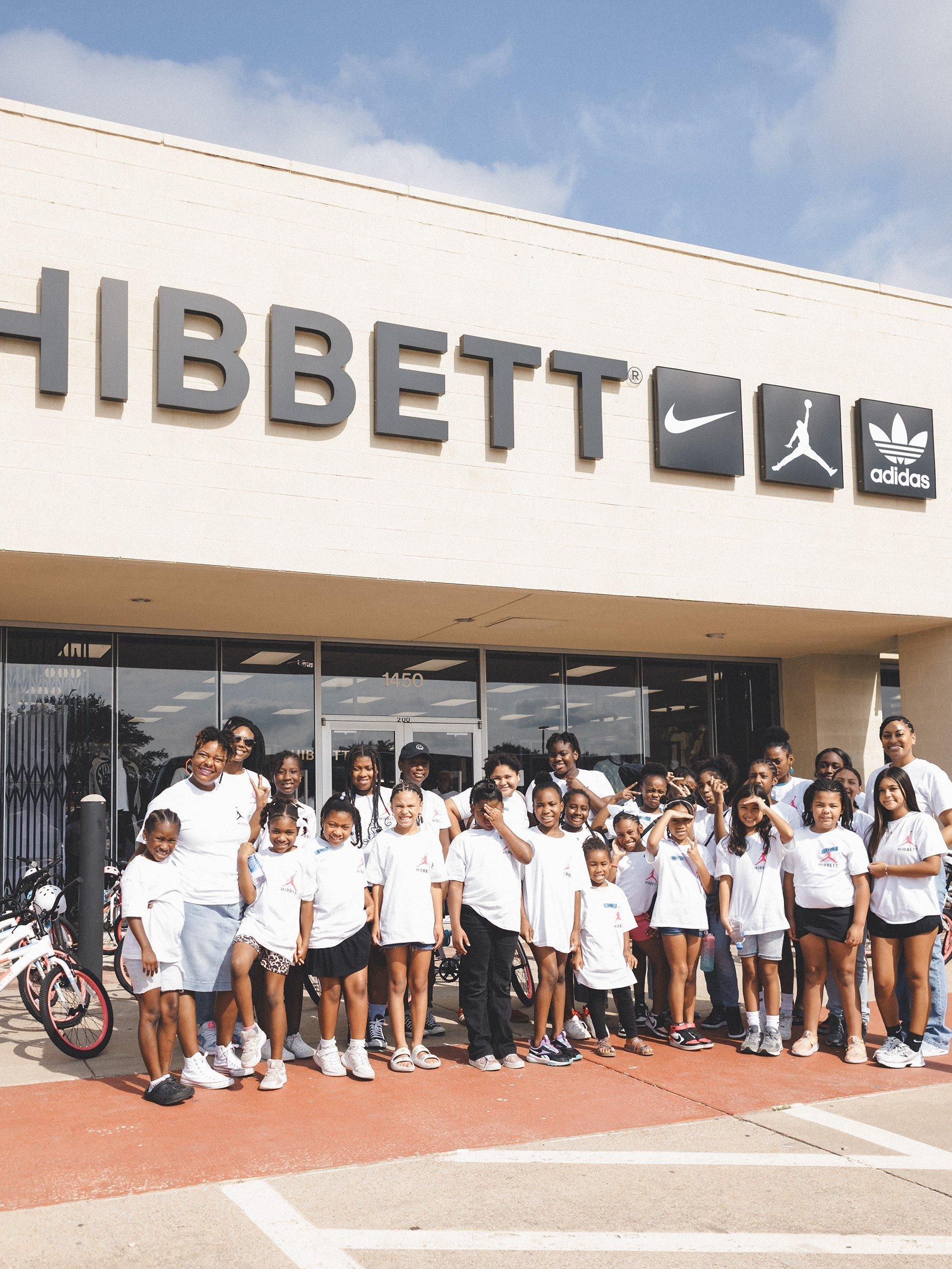 A large group of children and adults, all wearing white t-shirts, stands in front of a Hibbett store for a group photo. The store's sign includes the logos for Hibbett, Nike, Jordan, and adidas.
