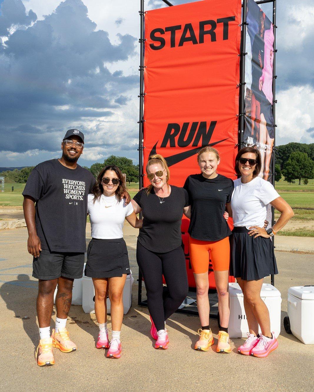 A group of five people, four women and one man, stand smiling in front of a large red banner with "START RUN" written on it. They are all wearing running or athletic clothing, and some are wearing brightly colored Nike running shoes. The background is a grassy field with trees and a cloudy sky.