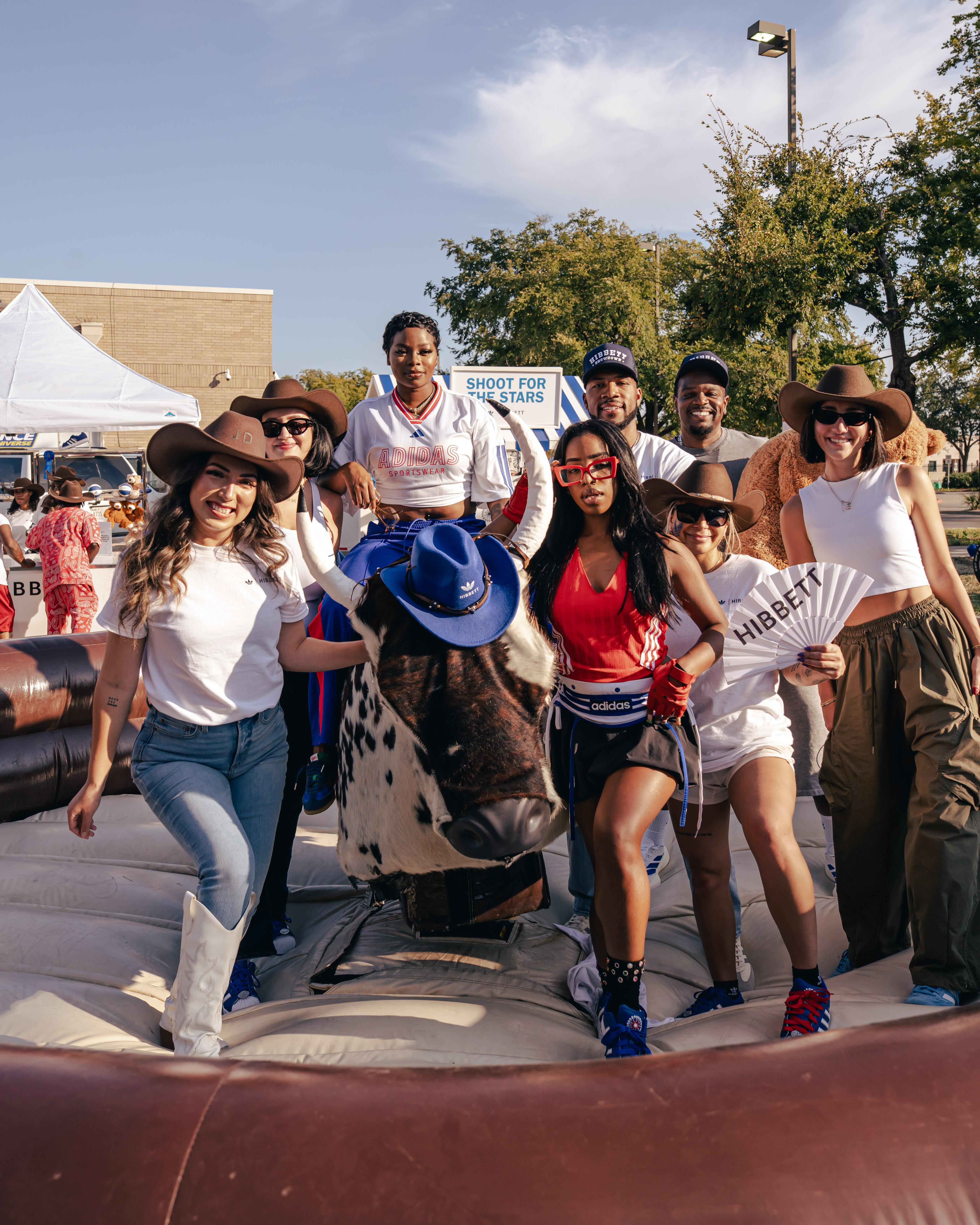 A group of people poses for a picture on an inflatable mechanical bull. Many are wearing cowboy hats and casual outfits. One woman in the center holds up a fan with "HIBBETT" written on it.