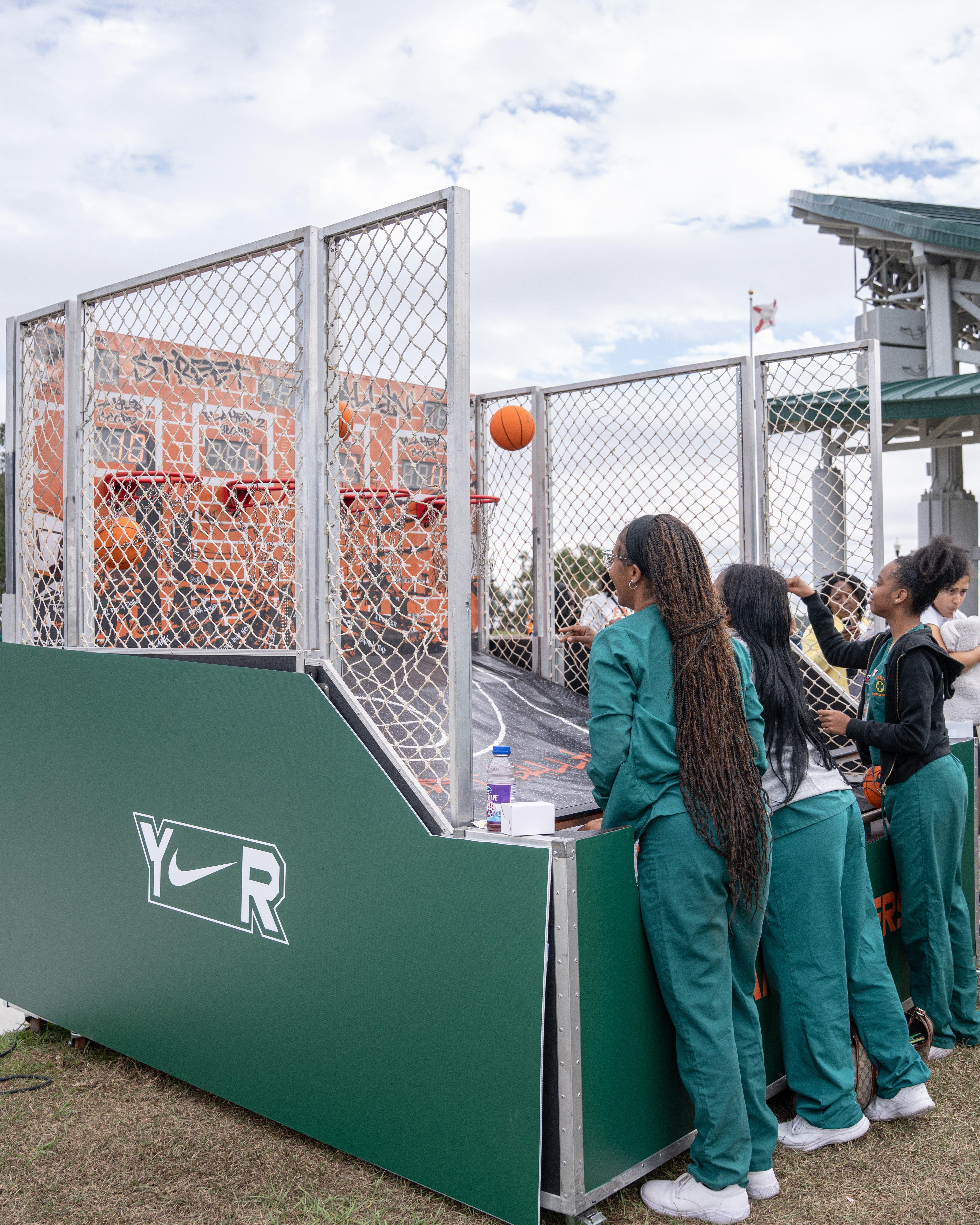 A young woman wearing a black t-shirt and camouflage pants is playing mini-golf on a portable green with a flag that reads HIBBETT. Other people are visible in the grassy background.