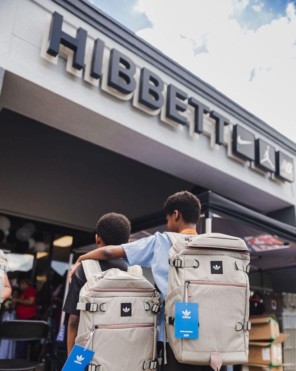 Two children wearing adidas backpacks outside a Hibbett store entrance.