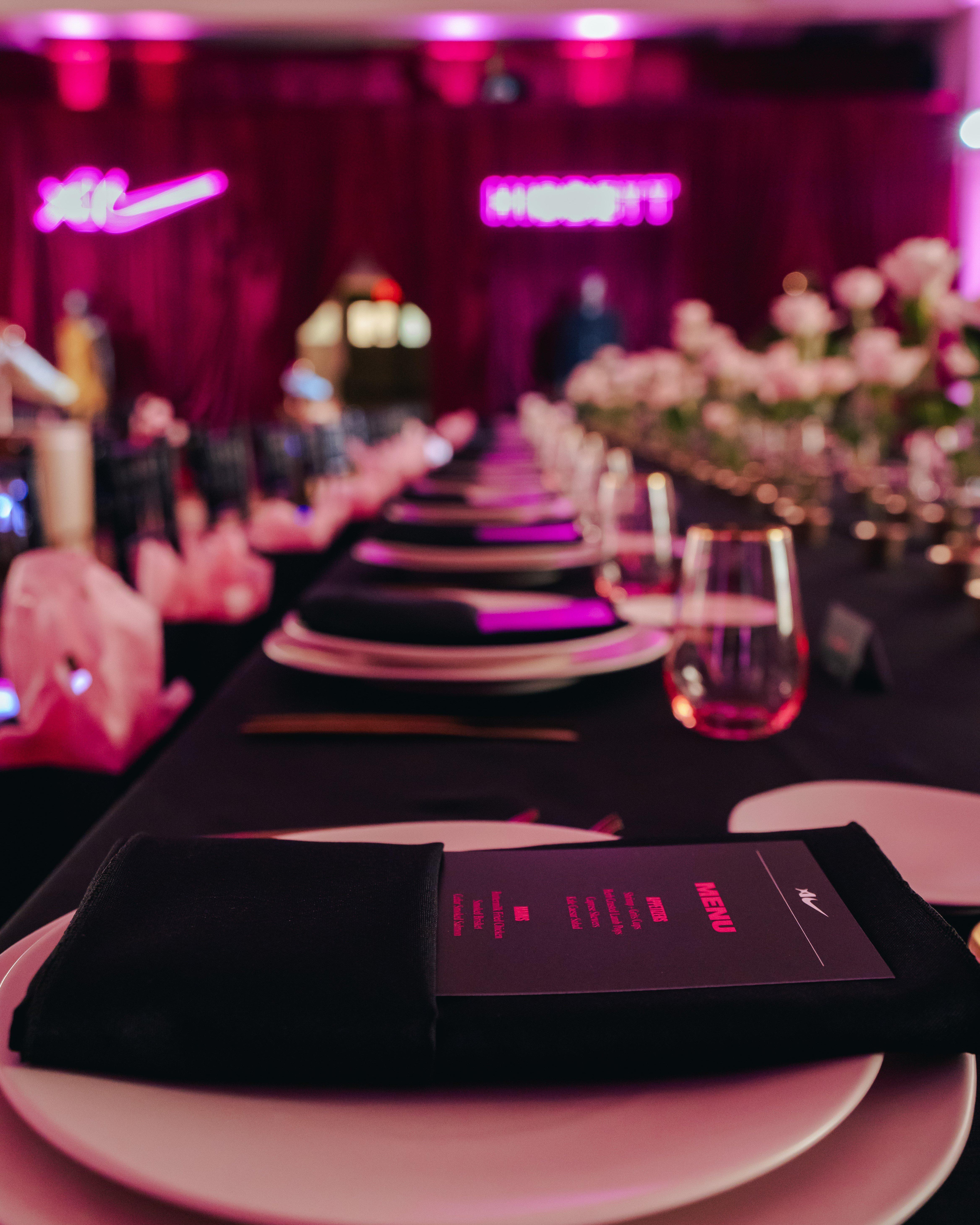 A long formal dinner table set with black tablecloths, white plates, and black napkins at a Hibbett and Nike event. In the background, vibrant pink neon signs displaying the Nike Swoosh and Hibbett logo illuminate the room alongside floral arrangements.