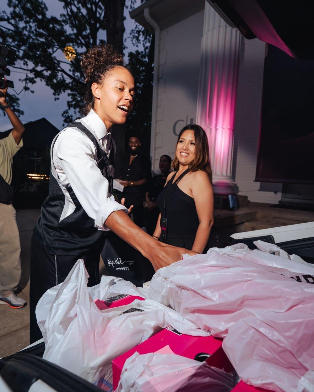A woman with her hair in a bun reaches into the trunk of a vehicle filled with hot pink A'ja Wilson shoe boxes and plastic bags during a Hibbett and Nike event. A smiling woman in a black halter top looks on in the background outside a building with pink pillar lighting.