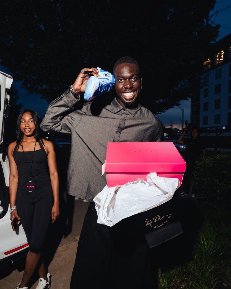 A smiling man holds a bright blue Nike sneaker up to his ear like a phone while carrying a hot pink A'ja Wilson shoe box and a black Hibbett shopping bag outdoors. A female staff member in a black outfit stands in the background next to a white vehicle at night.